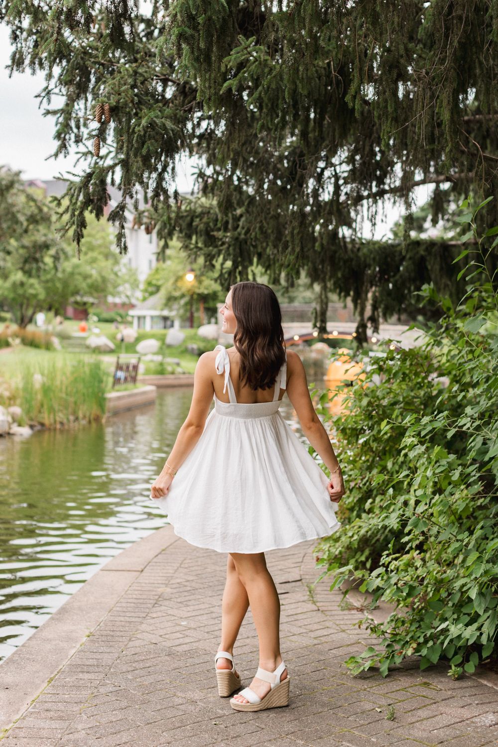 A woman in a flowy white dress twirls on a pathway beside a pond under hanging willow branches.