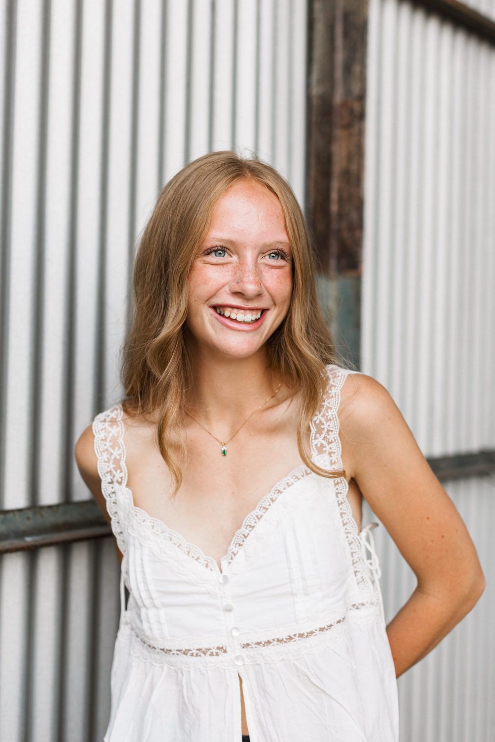 Young woman in white lace dress poses and smiles against a corrugated metal wall.
