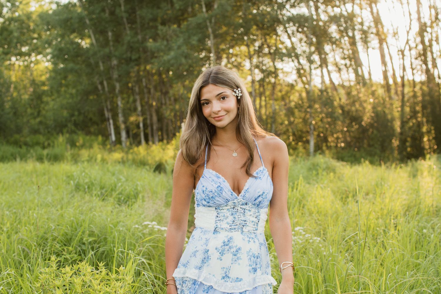 Young woman in blue floral sundress with flower hair accessory smiling in sunlit green meadow with trees.