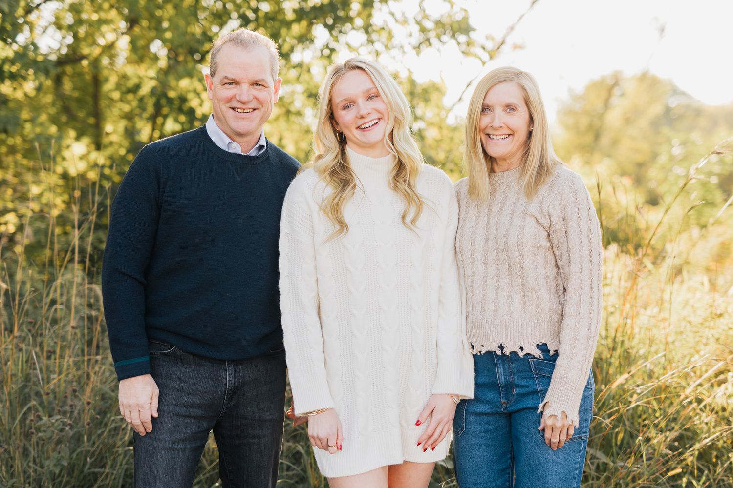 Three people in warm sweaters pose together outdoors during golden hour.