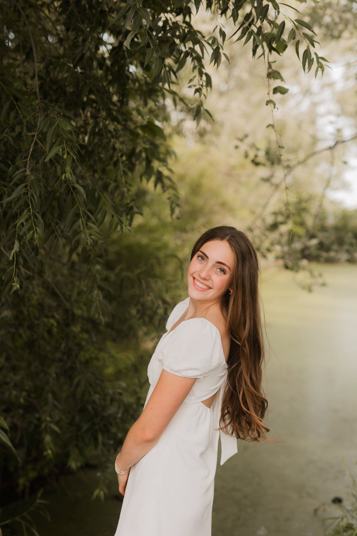Brunette woman in white off-shoulder dress smiling by a willow tree near a pond.