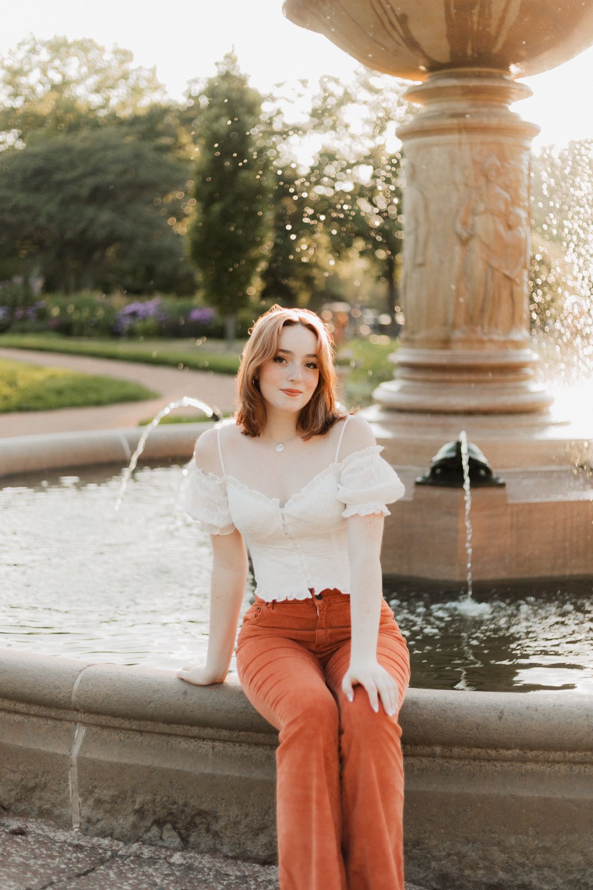 Young woman in white crop top and rust pants sits on ornate stone fountain in sunlit park.