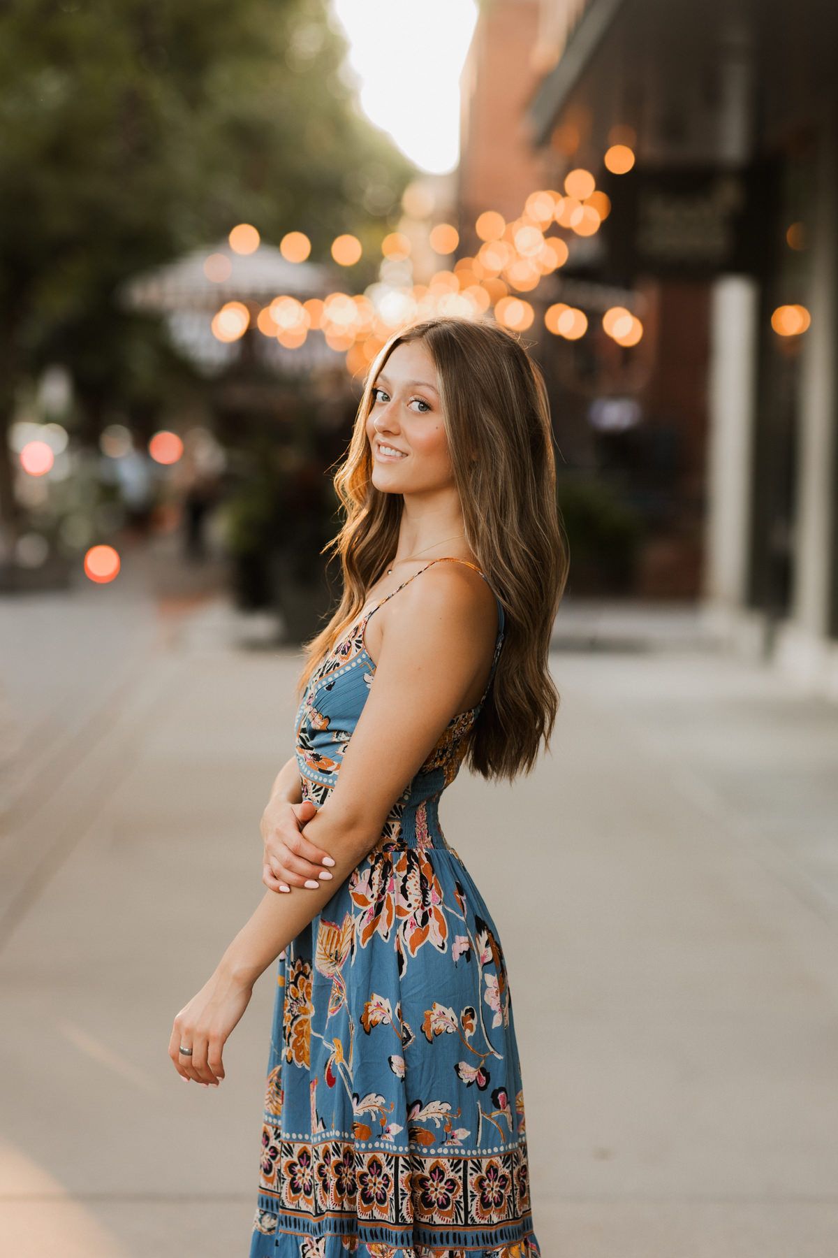 Young woman in floral blue dress smiles over shoulder on a charming city street at sunset.