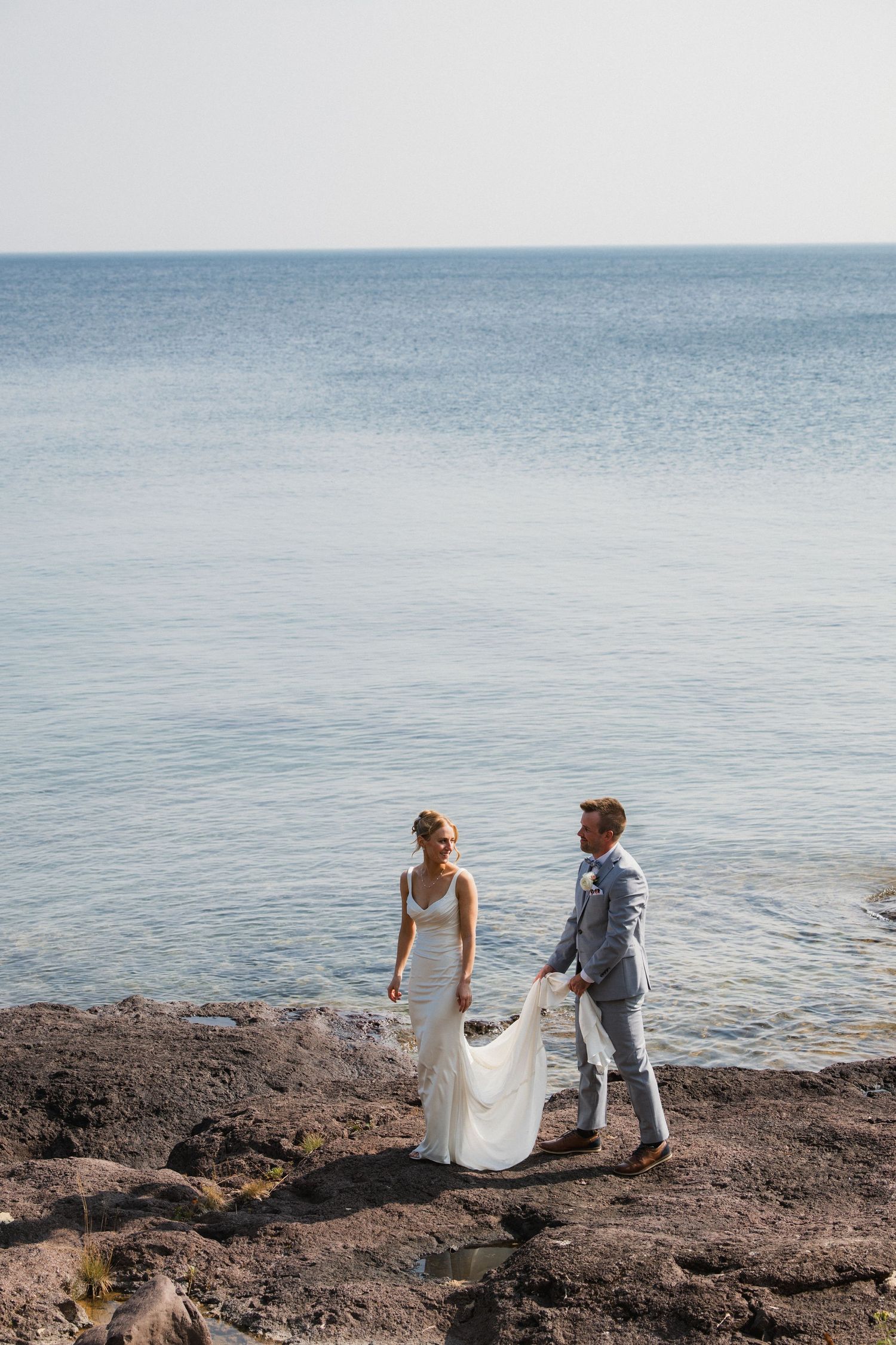 Bride and groom stand on rocky shoreline, groom holding wedding dress train, calm blue ocean backdrop.