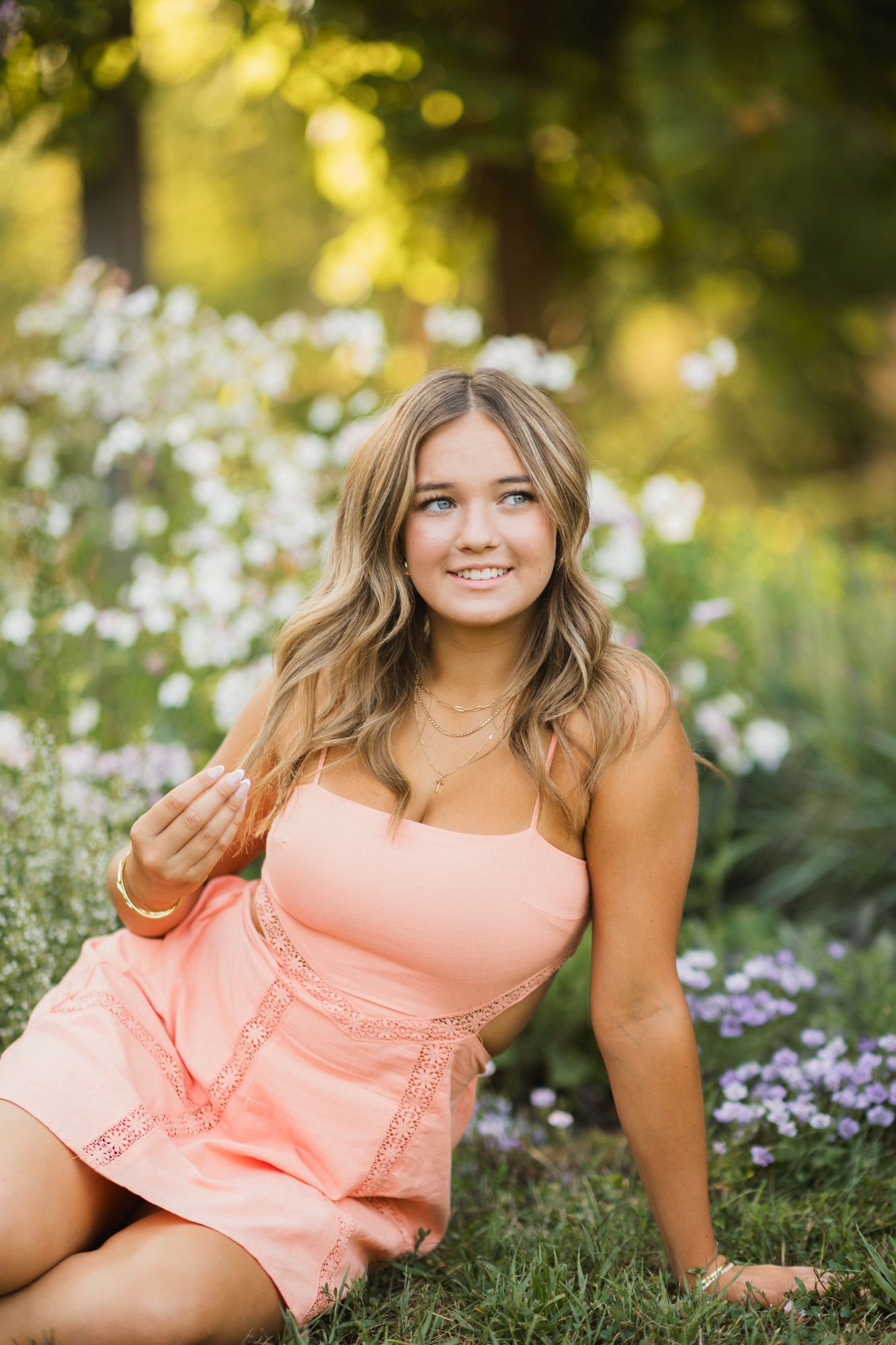 Young woman in peach sundress sitting among wildflowers in a sunlit garden, smiling warmly.