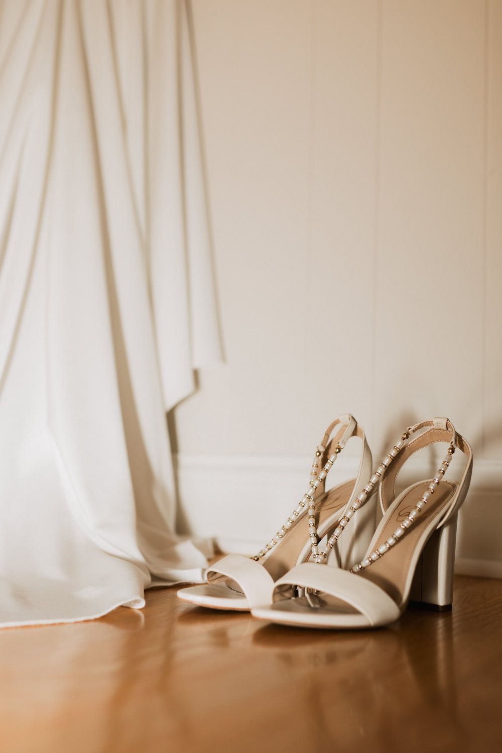 White wedding shoes peek out from beneath a flowing dress hem on a wooden floor.