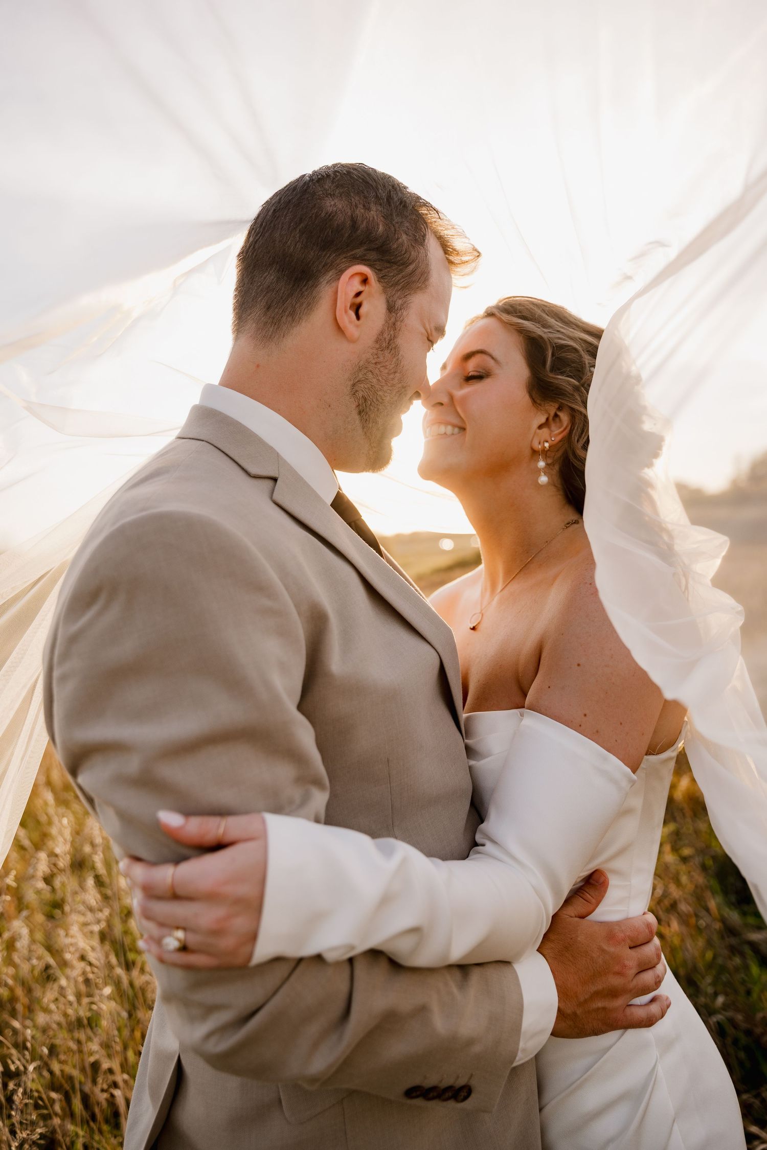 A bride and groom embrace tenderly under a flowing veil at golden hour in an outdoor field setting.