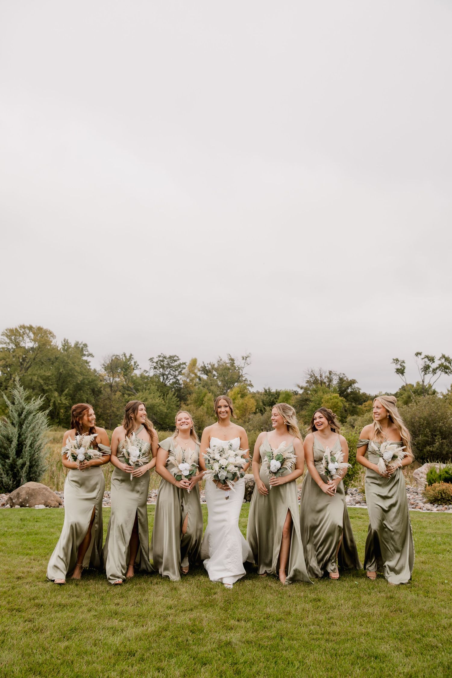 Bride in white gown poses with six bridesmaids in sage green satin dresses holding white bouquets on lush green lawn.