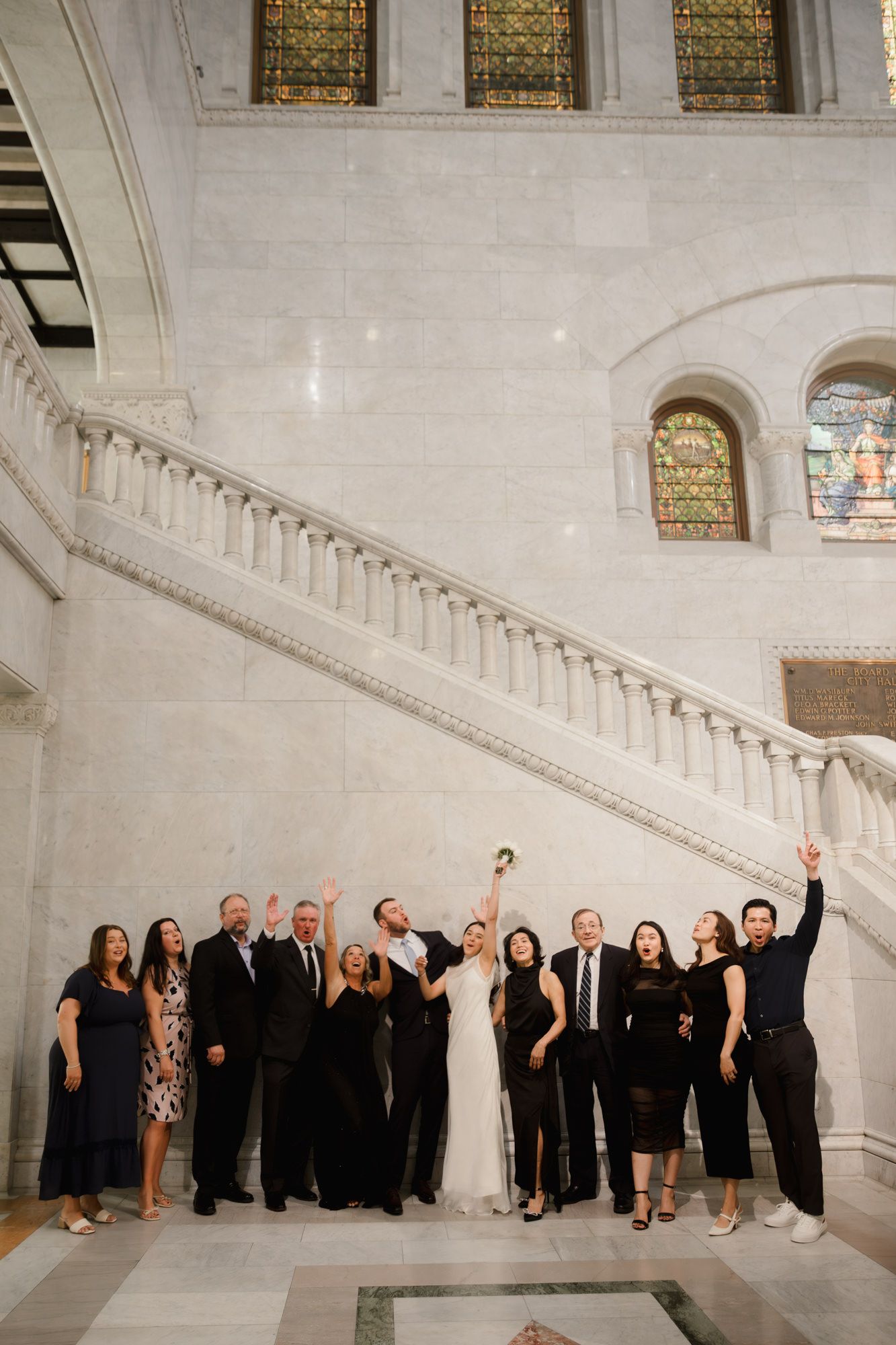 Wedding party celebrating in grand marble hall with staircase and stained glass windows, bride tossing bouquet.