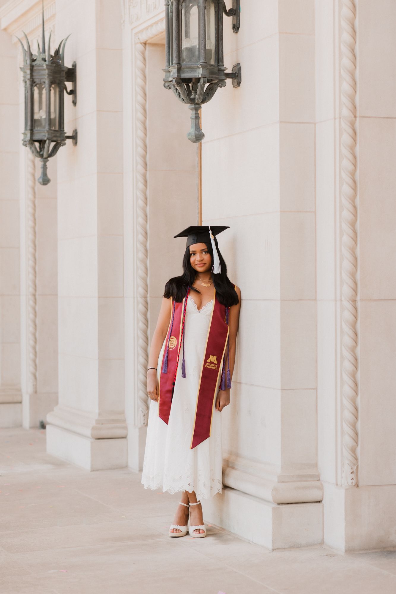 A graduate in white dress, maroon stole, and cap poses by ornate columns with vintage lanterns.