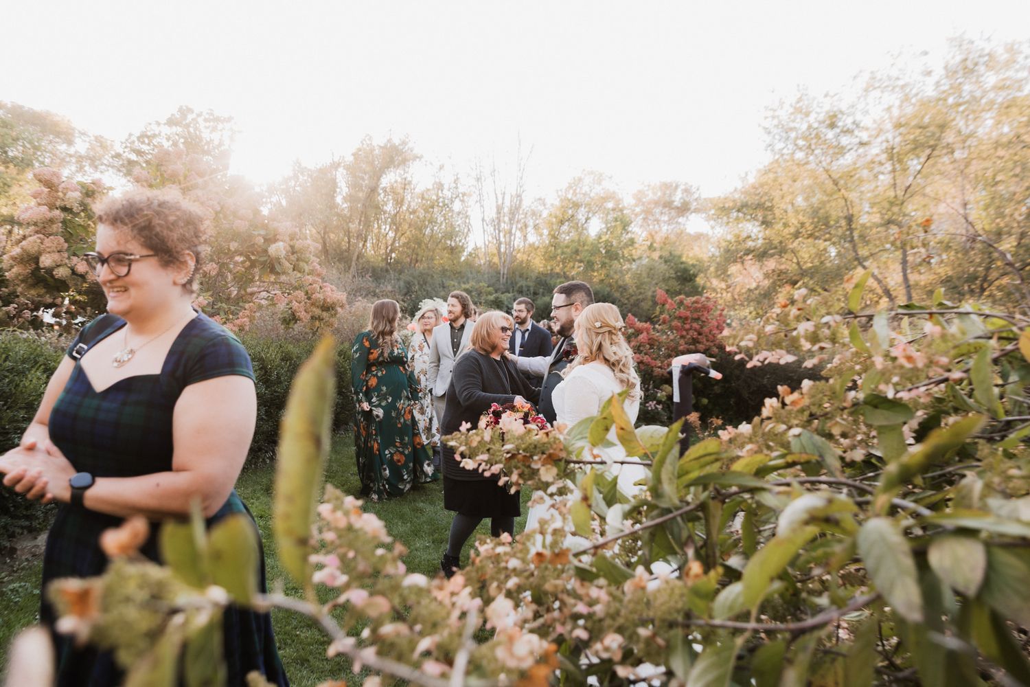 Guests gather outdoors among blooming shrubs during a garden wedding ceremony in warm autumn sunlight.