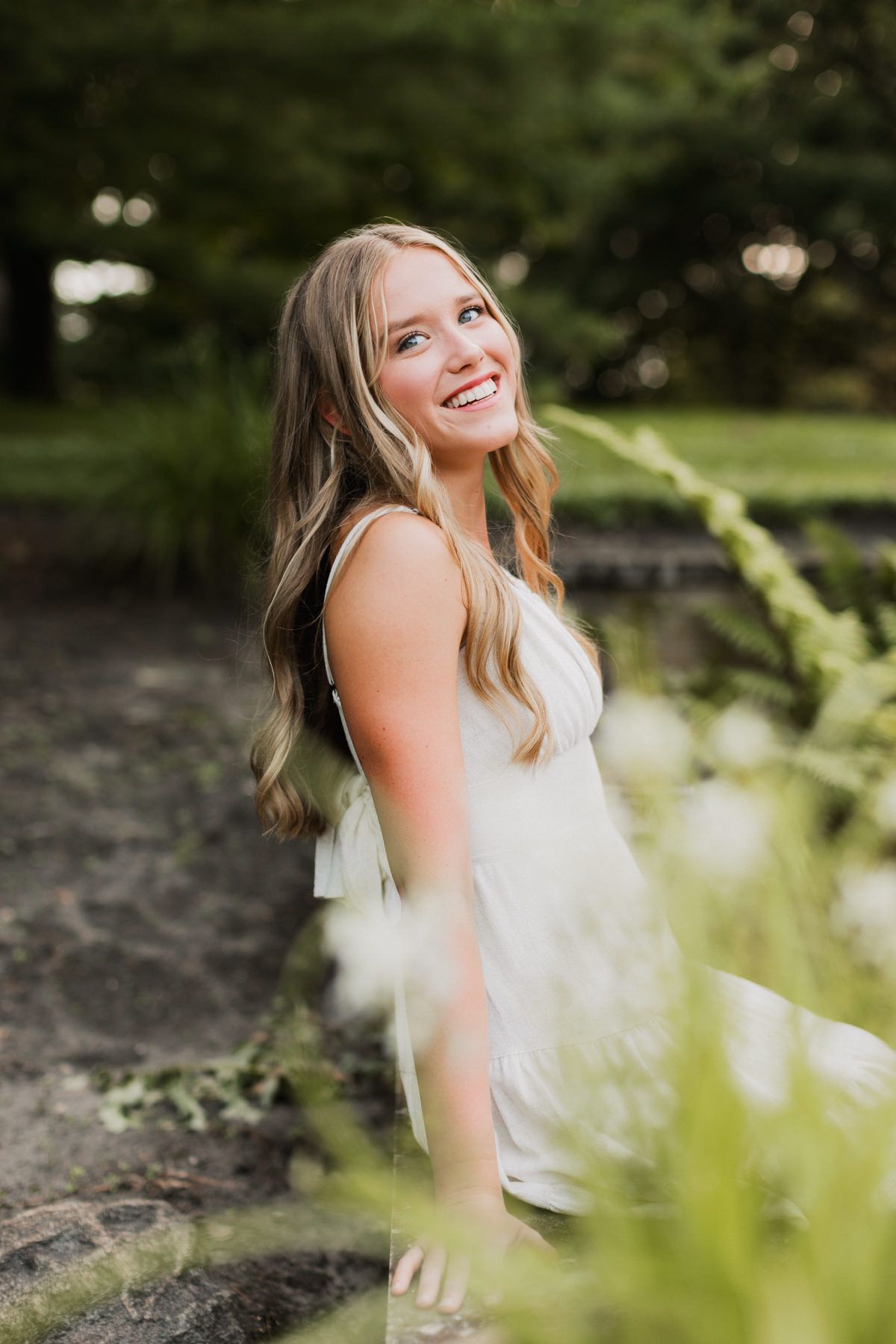 Laughing blonde woman in white dress sitting on rocks surrounded by lush green garden.