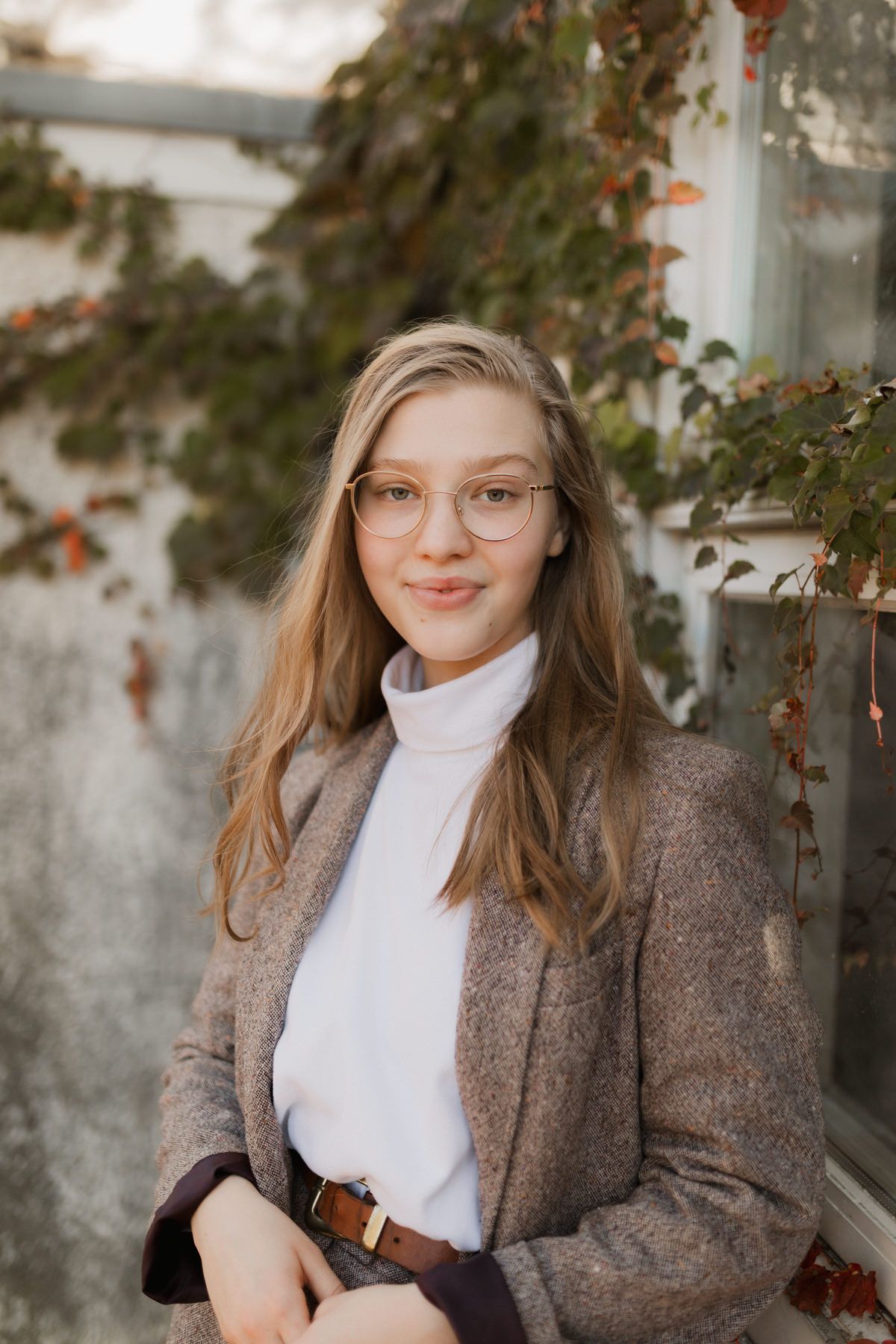 Young blonde woman wearing glasses and tweed blazer with white turtleneck posing near autumn vines.