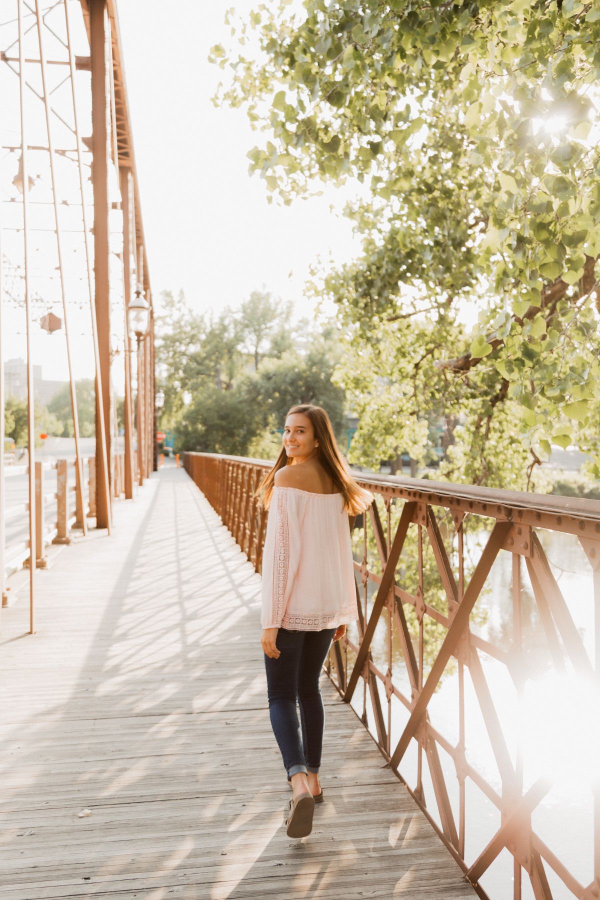 Young woman in pink off-shoulder top walking on rustic iron bridge surrounded by sunlit green trees.