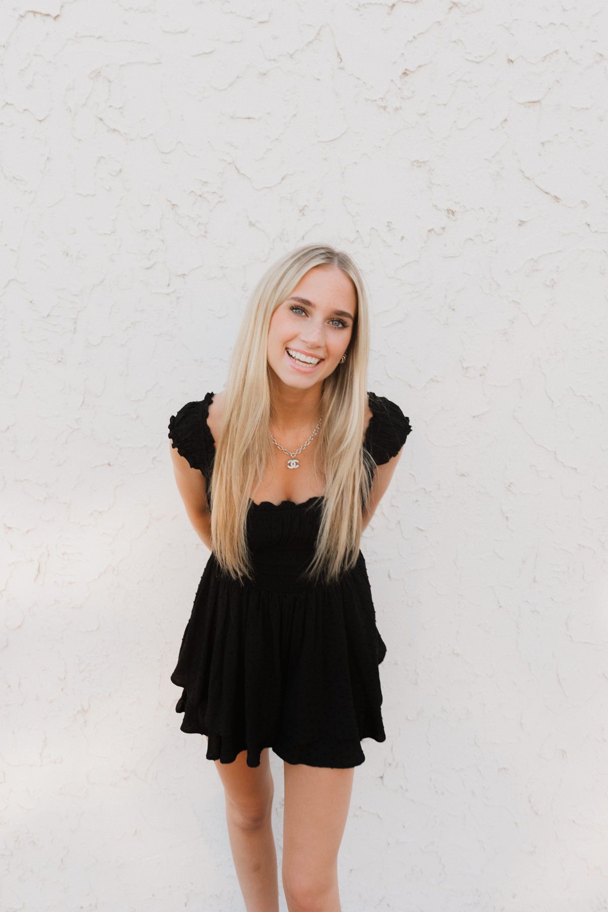 Smiling blonde woman in black ruffled mini dress posing against a white textured wall.