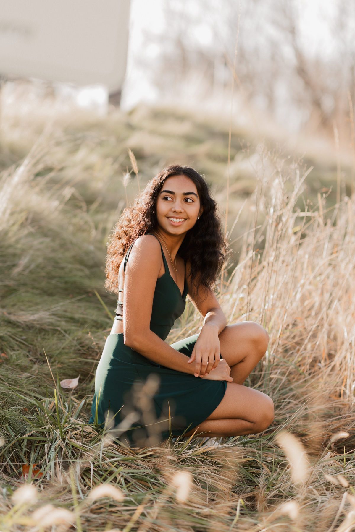 Young woman in green dress sitting in tall golden grass field, smiling outdoors during golden hour.
