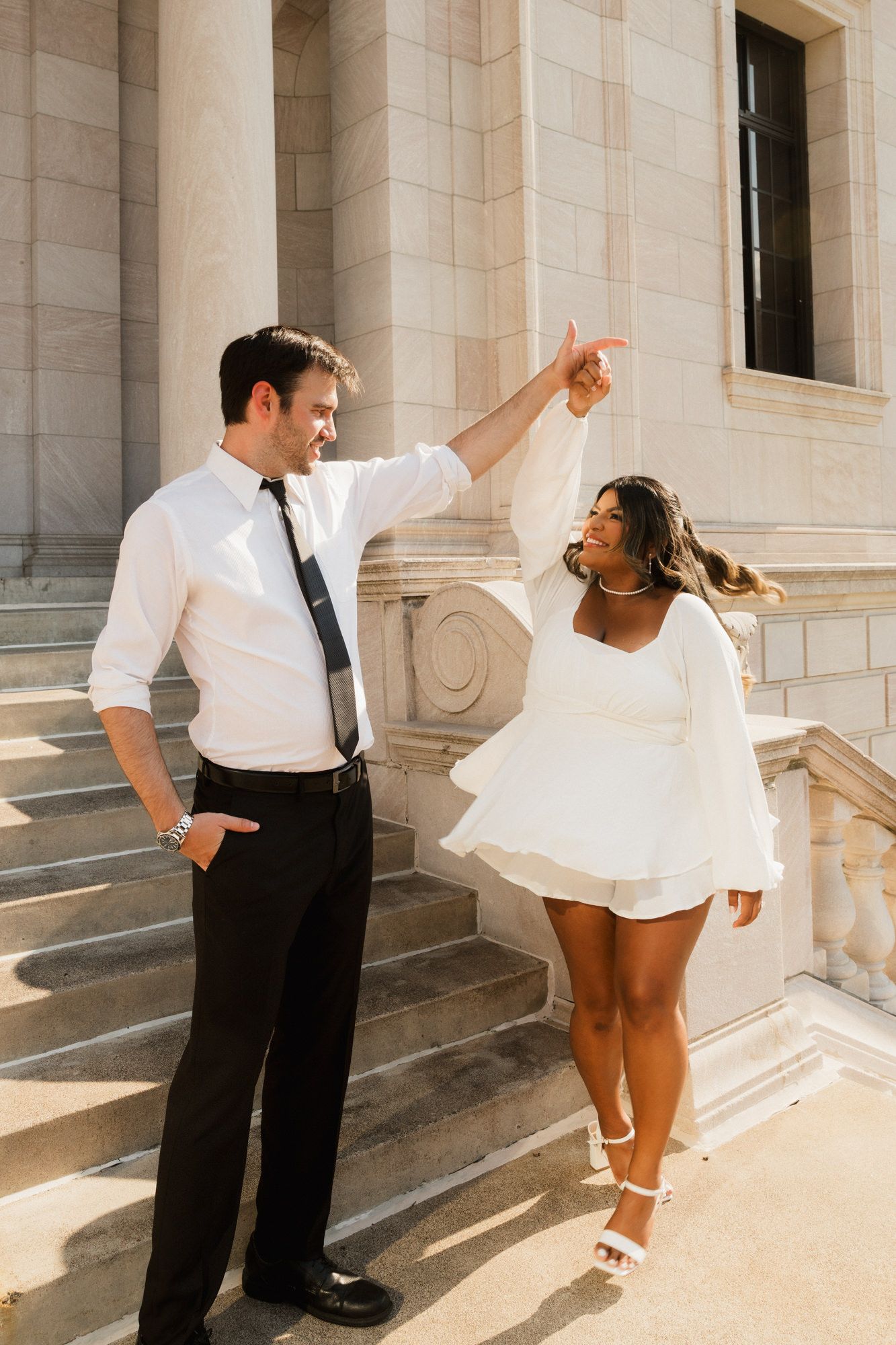 Couple dancing on stone steps, man in white shirt and tie spinning woman in white mini dress outdoors.