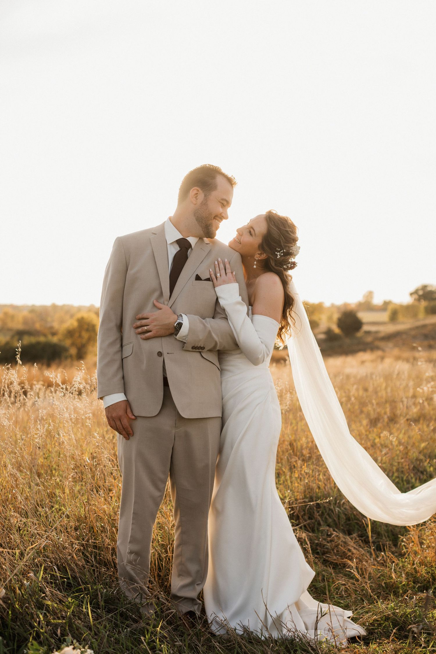 Smiling groom in a tan suit and bride in a white gown with a flowing veil embrace in a golden field at sunset.