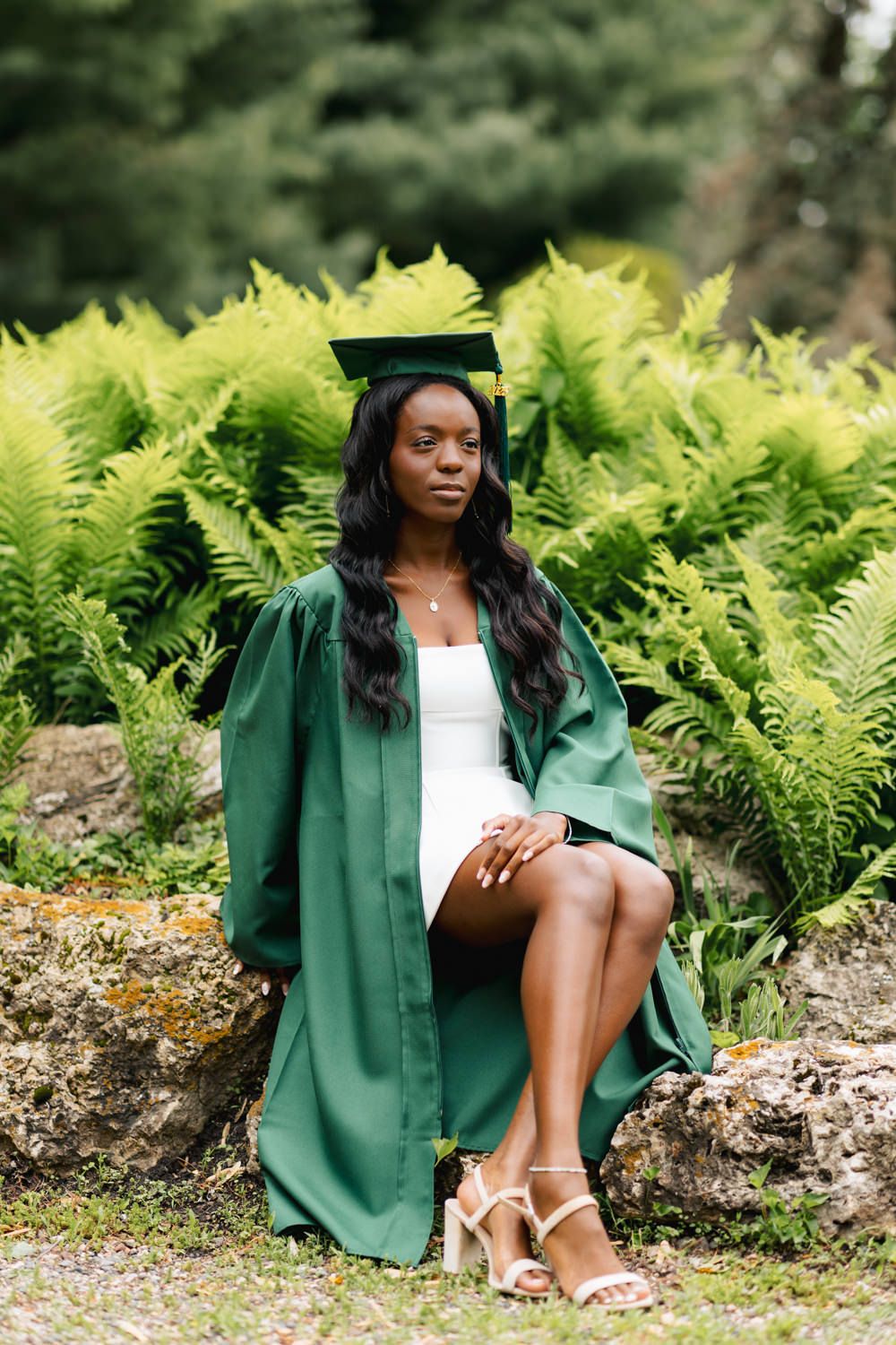 Graduate in green regalia poses elegantly on rock surrounded by lush ferns.