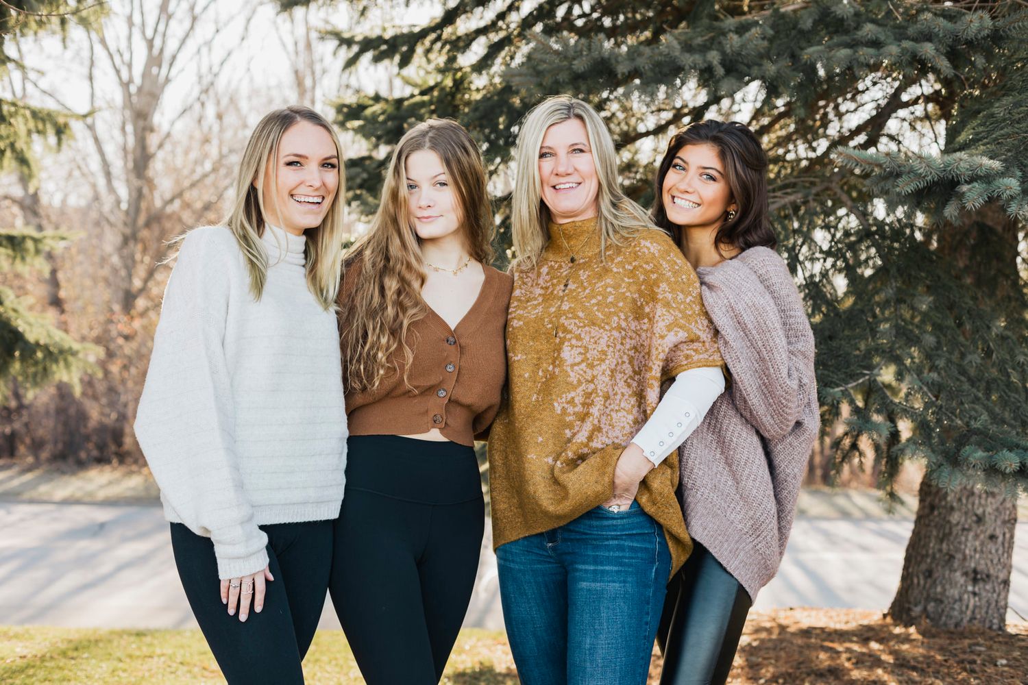 Four friends in stylish sweaters posing casually outdoors near evergreen trees.