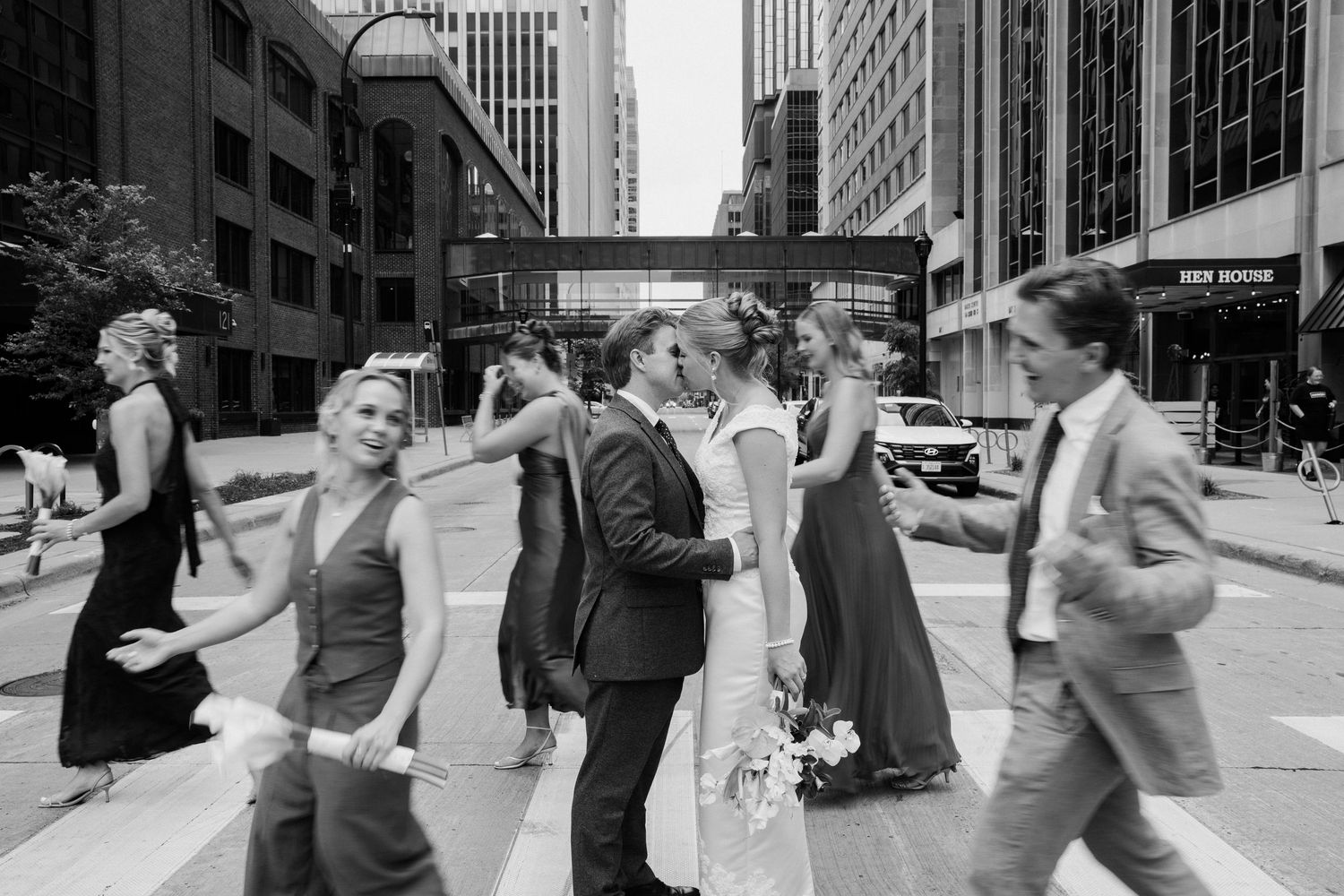 Black and white photo of a wedding party crossing a busy city street, bride and groom kissing in the center.