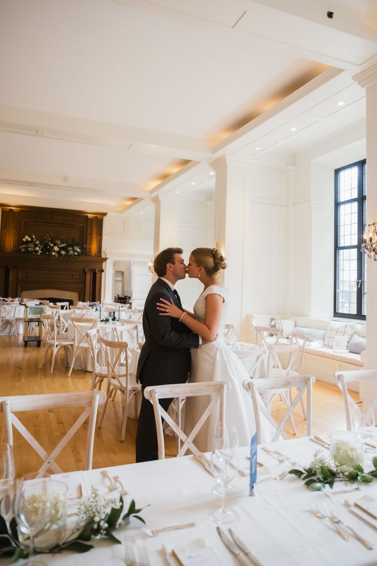 Bride and groom share a romantic kiss in an elegant white wedding reception hall with wooden floors.