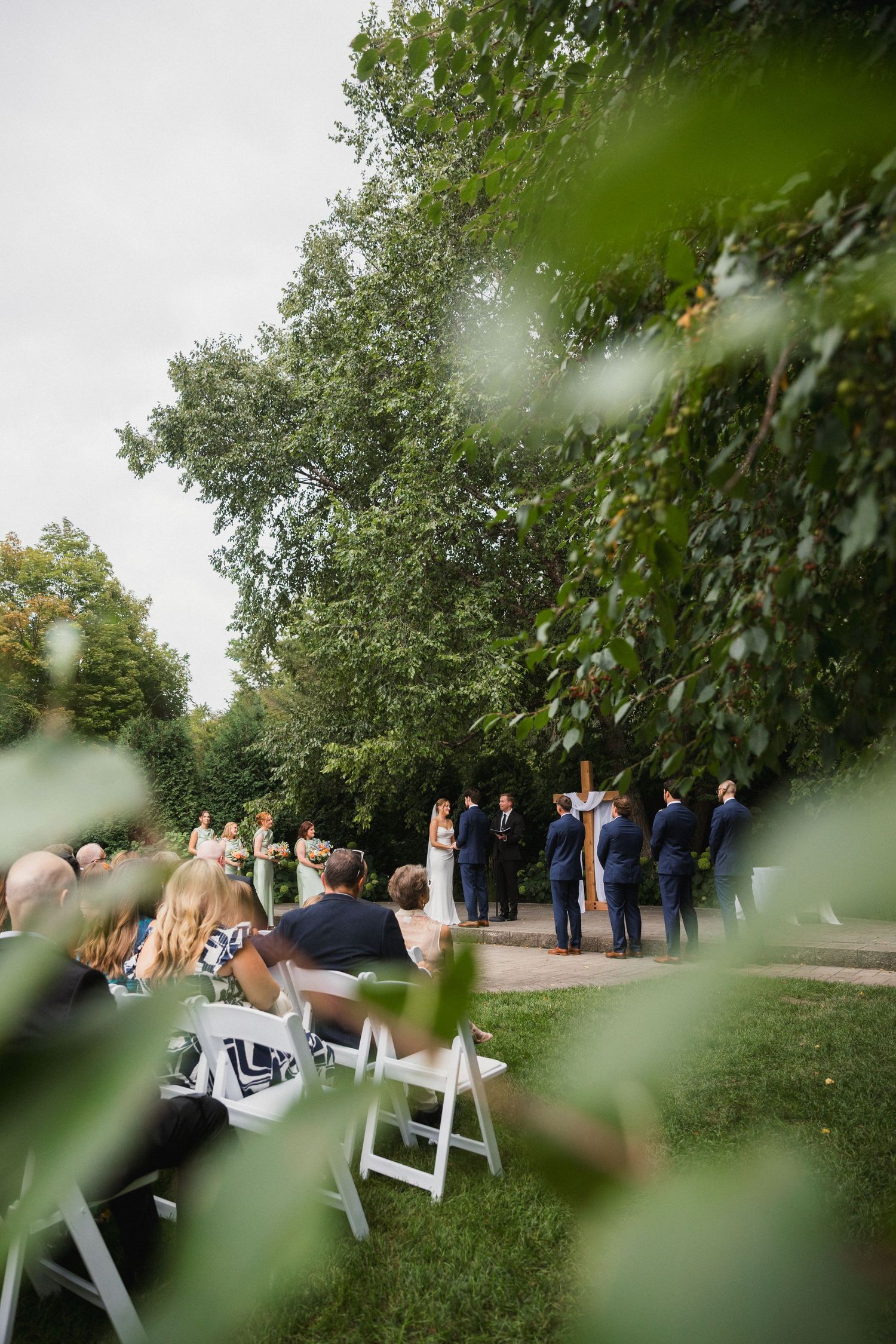 Outdoor wedding ceremony on green lawn with guests seated in white chairs, bridal party at altar surrounded by lush trees.
