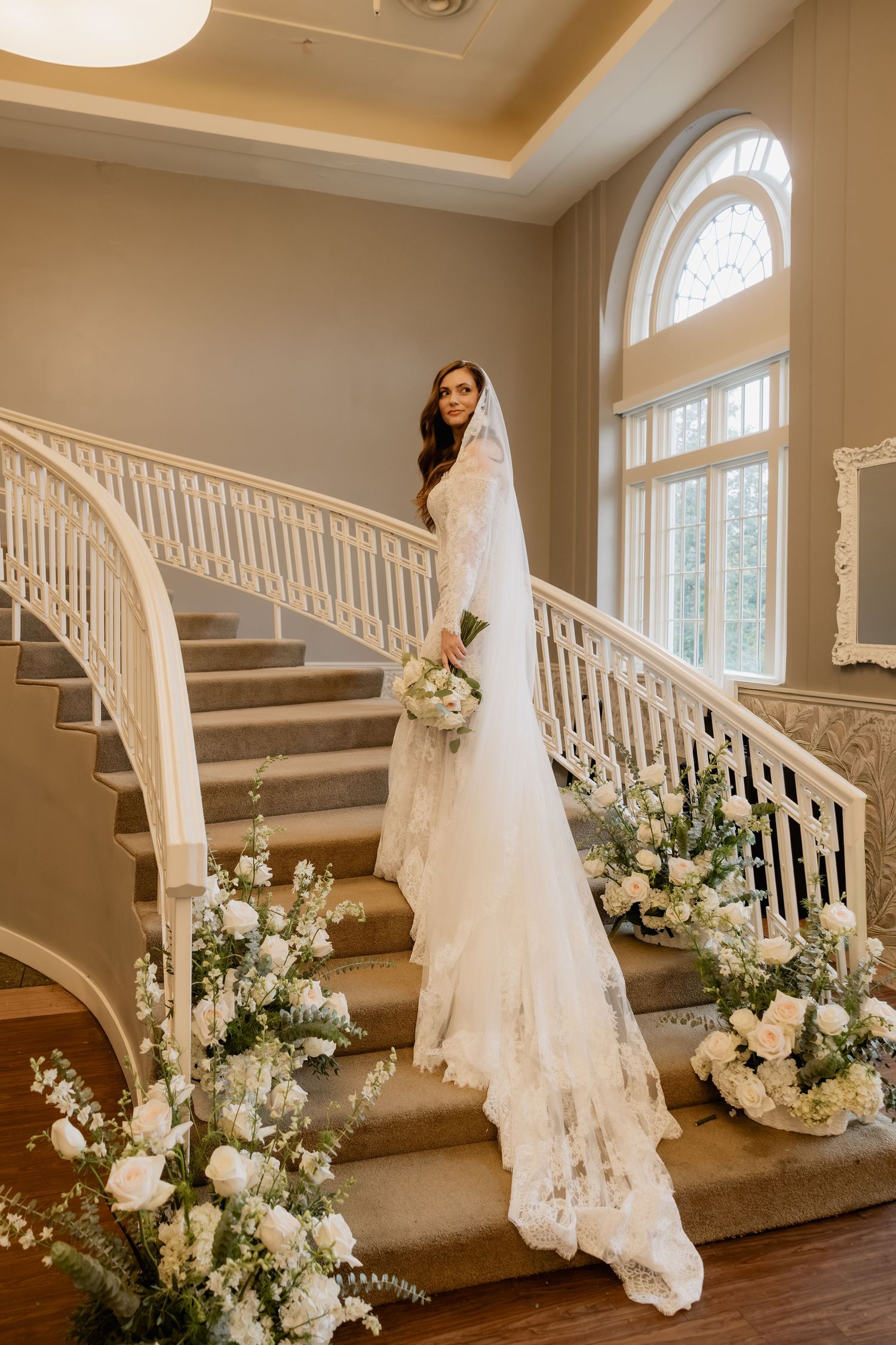 Bride in lace gown holding bouquet on elegant curved staircase adorned with white floral arrangements.