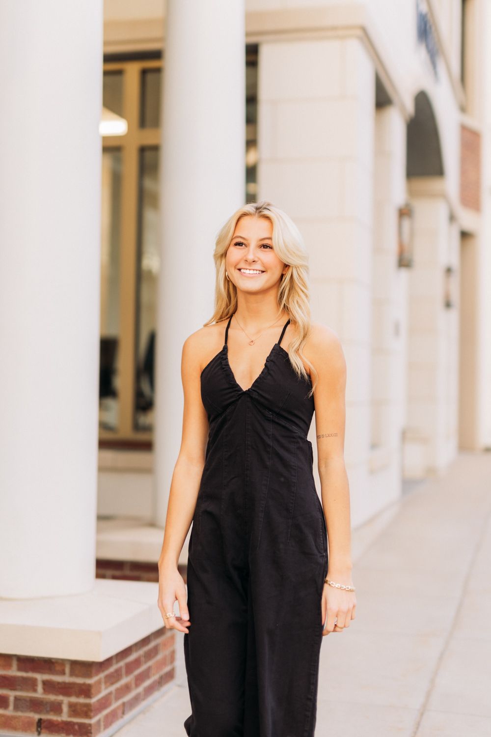 Woman in elegant black jumpsuit poses confidently against white pillars in an outdoor architectural setting.