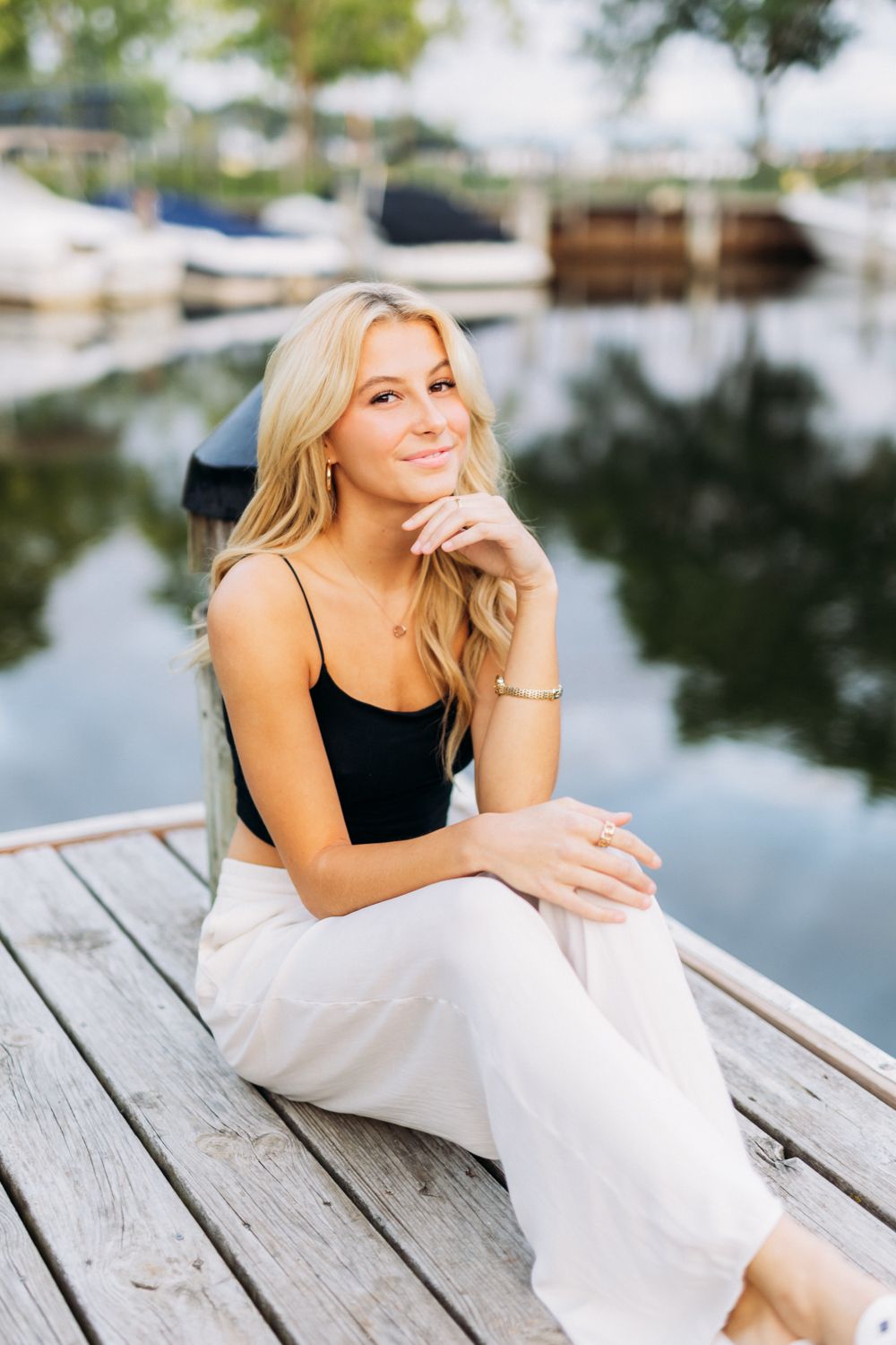 Someone wearing black and white sits casually on weathered dock planks with water and boats in background.