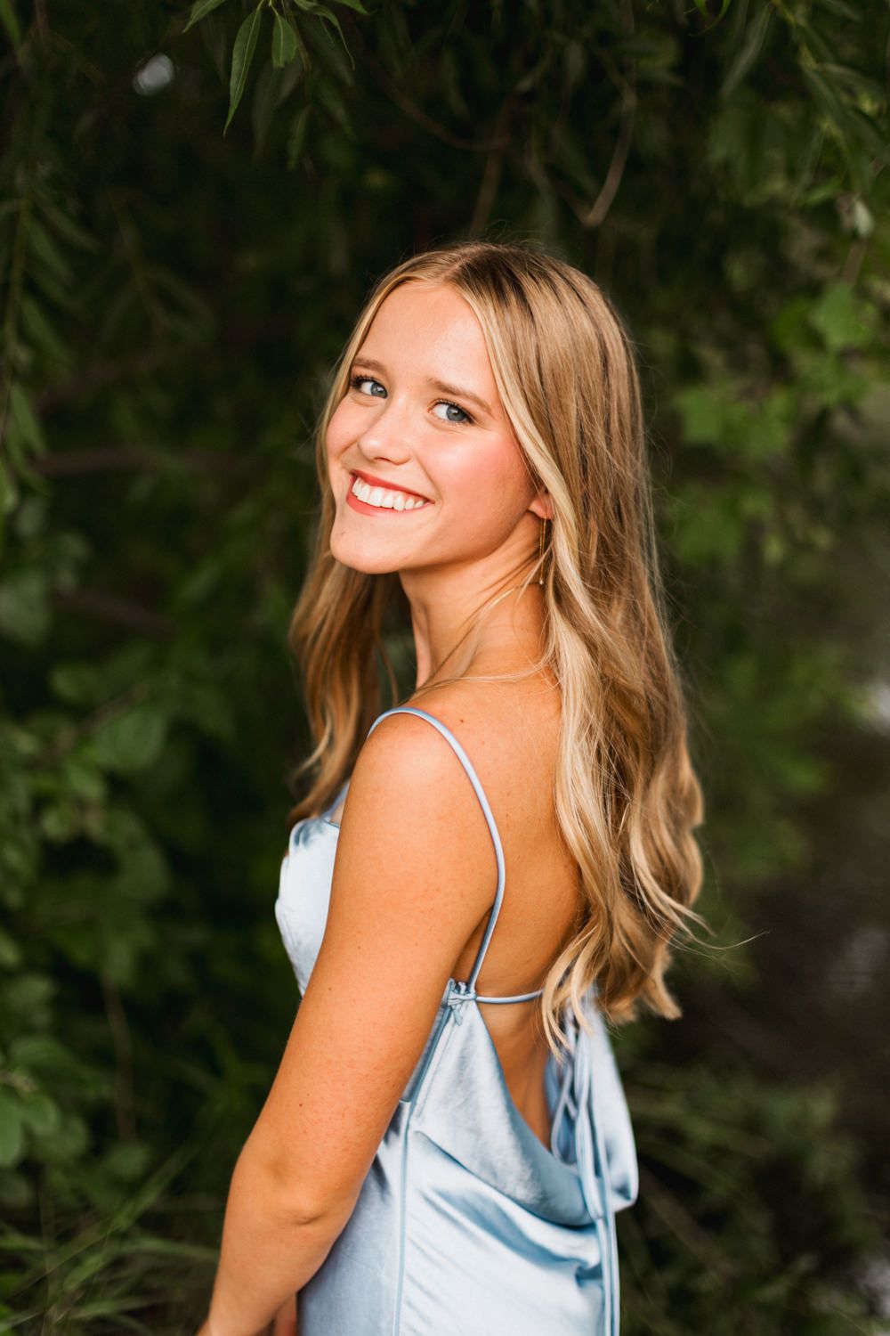 Woman in blue dress poses against green foliage background during outdoor portrait session.