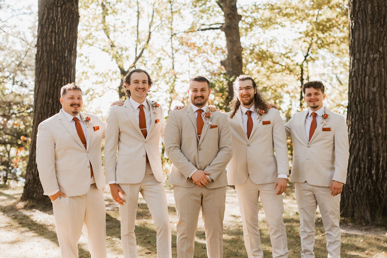Group in matching light colored suits and red ties posing together outdoors among trees.