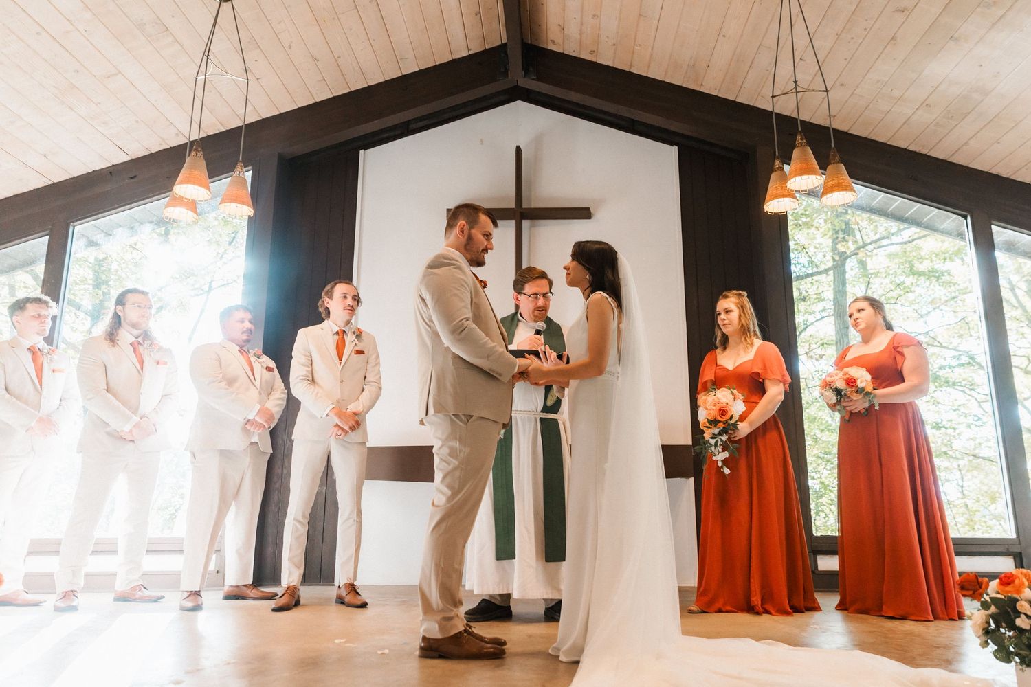 A wedding ceremony takes place in a bright chapel with wooden beams and hanging pendant lights.