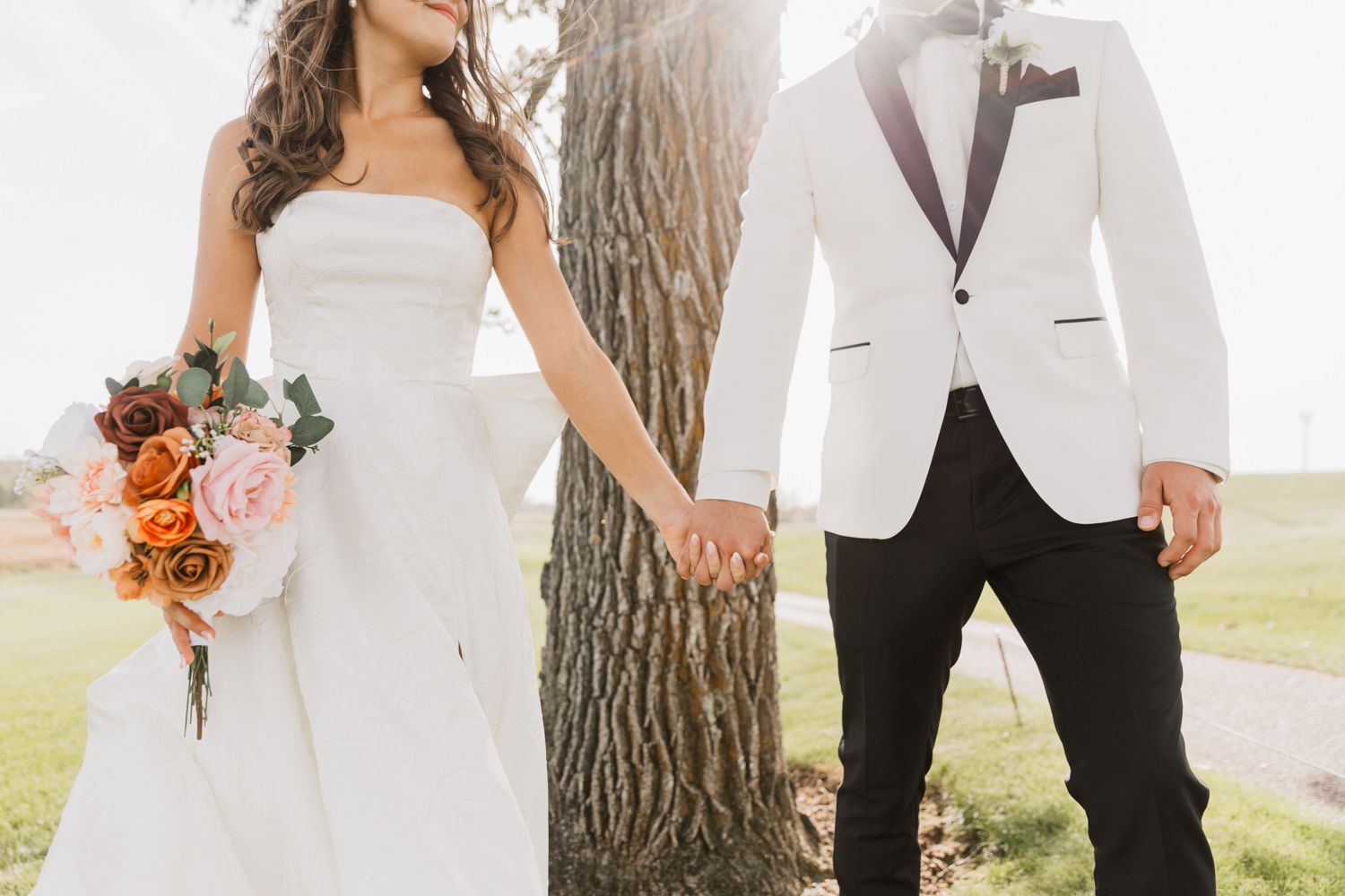 A wedding couple poses outdoors near a tree trunk, with the bride holding a vibrant orange and white bouquet.
