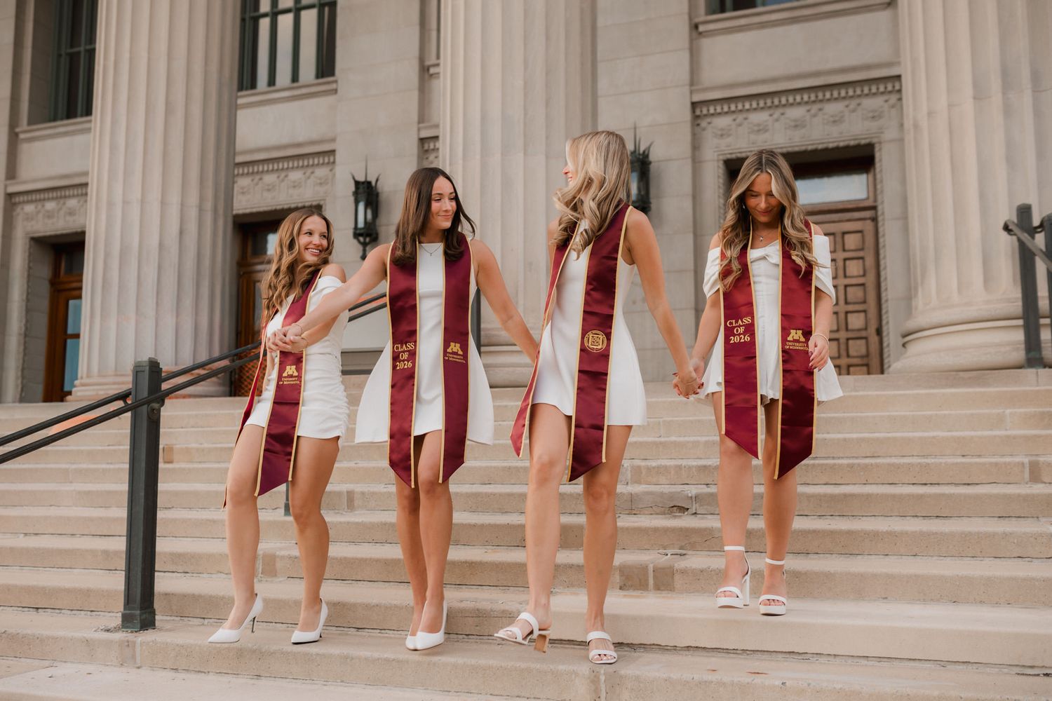 Four female graduates in white dresses and maroon graduation stoles hold hands on university building steps.
