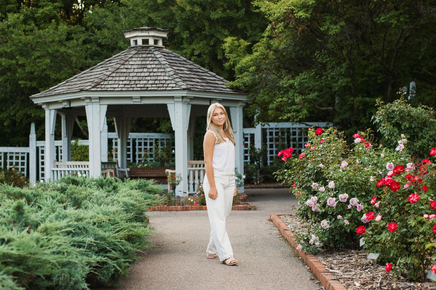 A woman in white outfit poses in a lush garden with a gazebo and blooming roses.