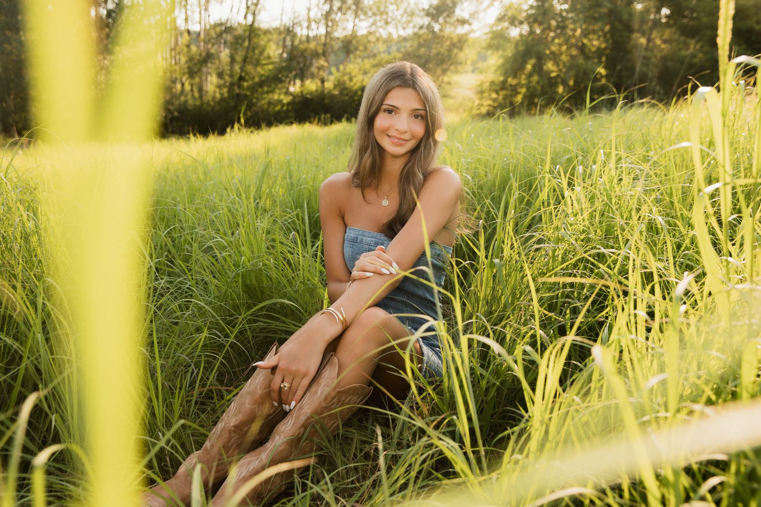Young woman smiling in a sunlit green meadow, sitting among tall grass in a casual denim outfit.