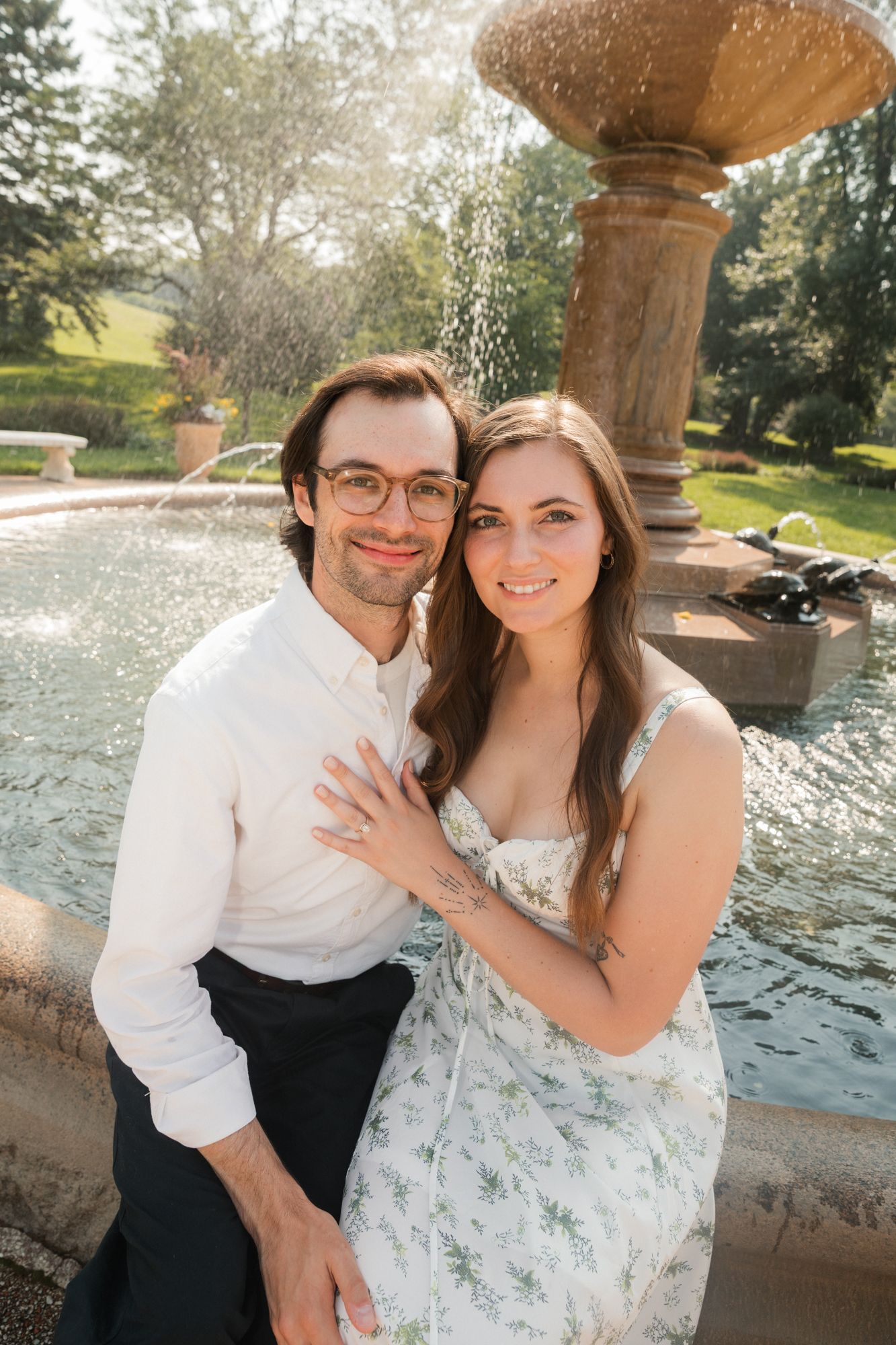 Smiling couple posing closely by a garden fountain, woman in floral sundress with hand on mans chest, sunny day.