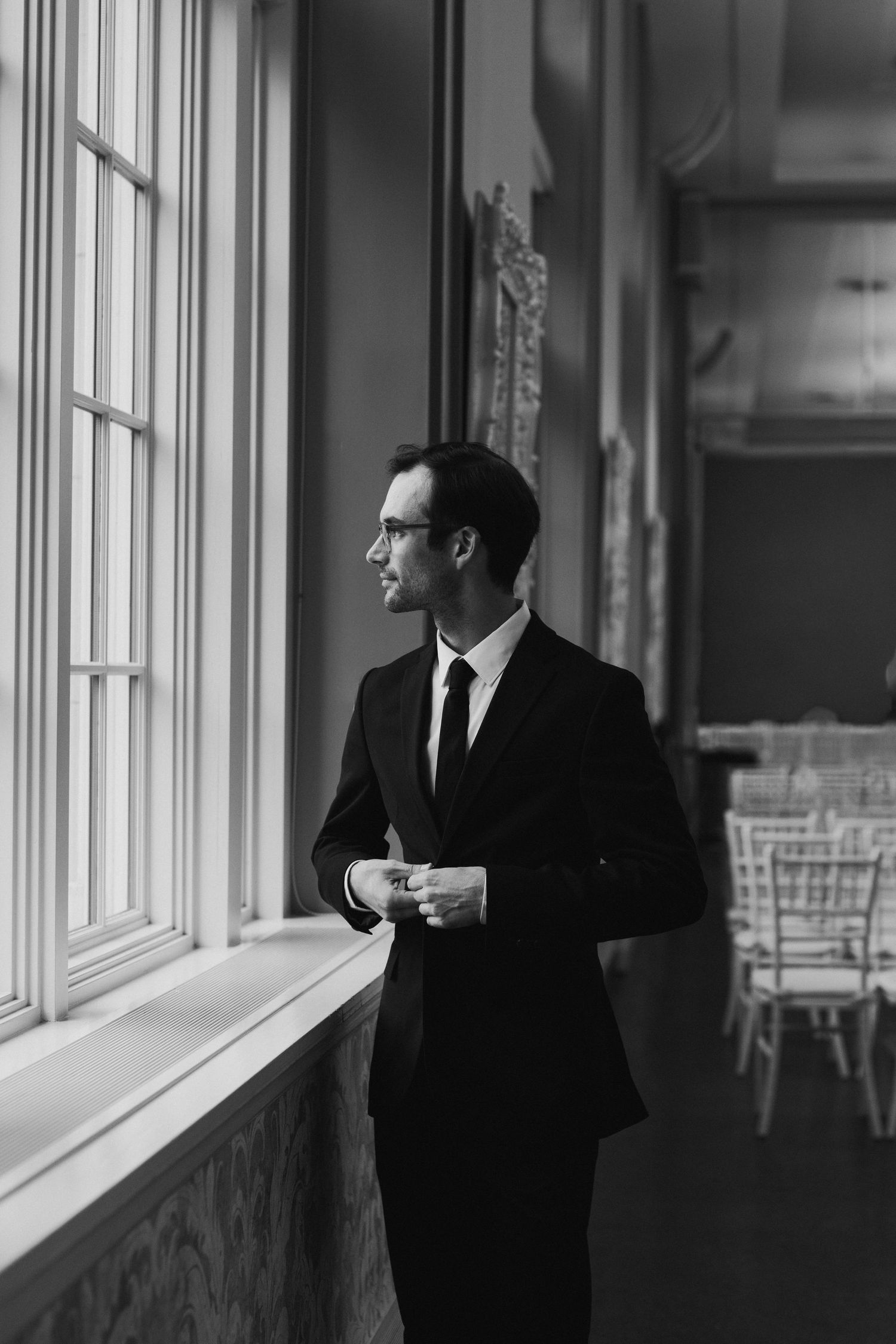 Black and white photo of a groom in a suit adjusting his cufflinks by a window before his wedding ceremony.