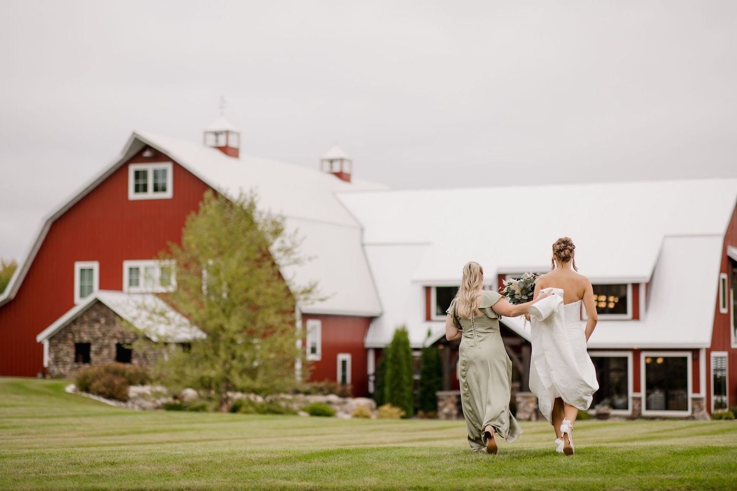 Bride and bridesmaids walking toward a red barn wedding venue on a lush green lawn.