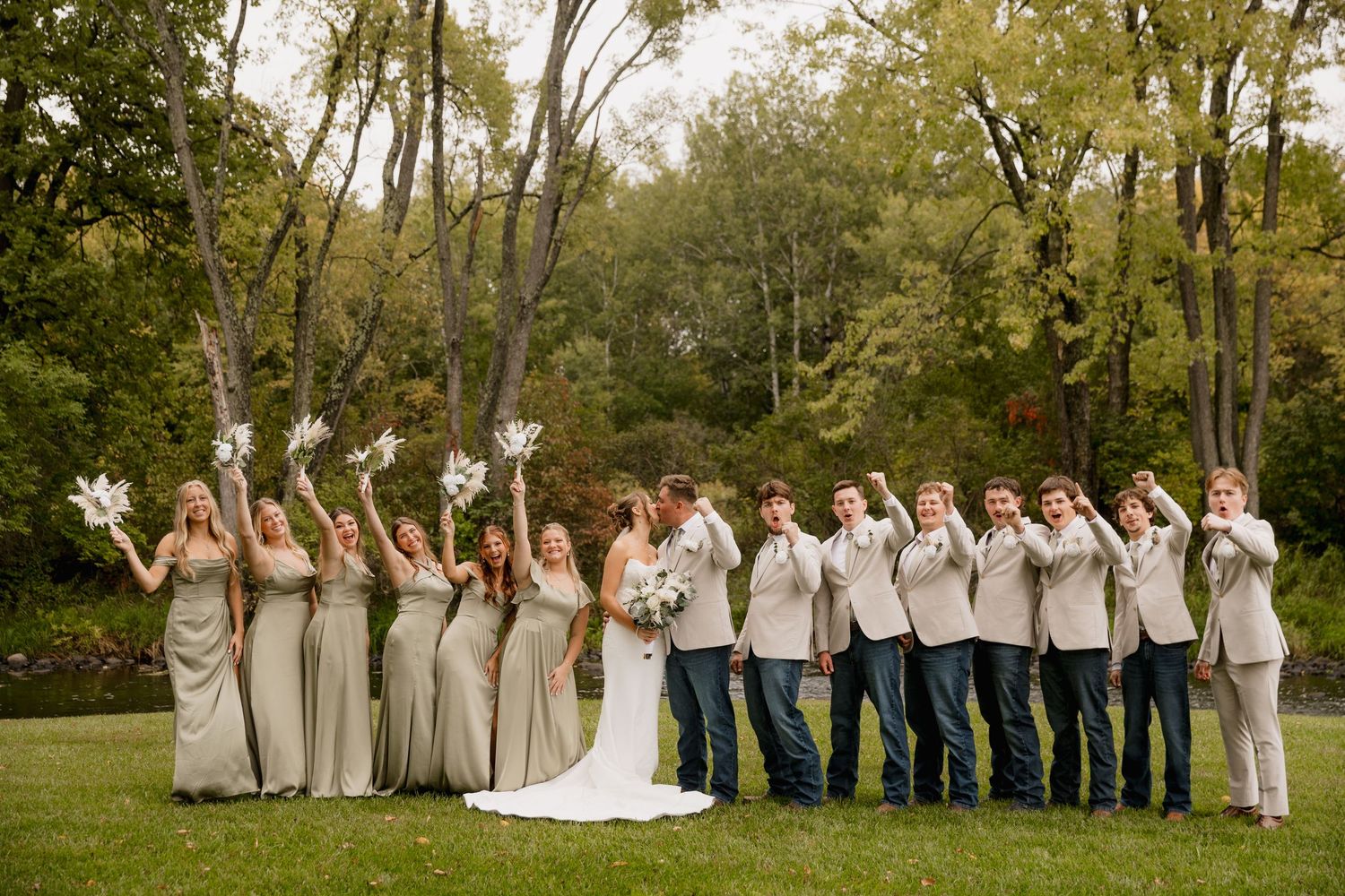 Wedding party in sage green dresses and gray suits poses outdoors, bridesmaids holding white pinwheels on lush green lawn.