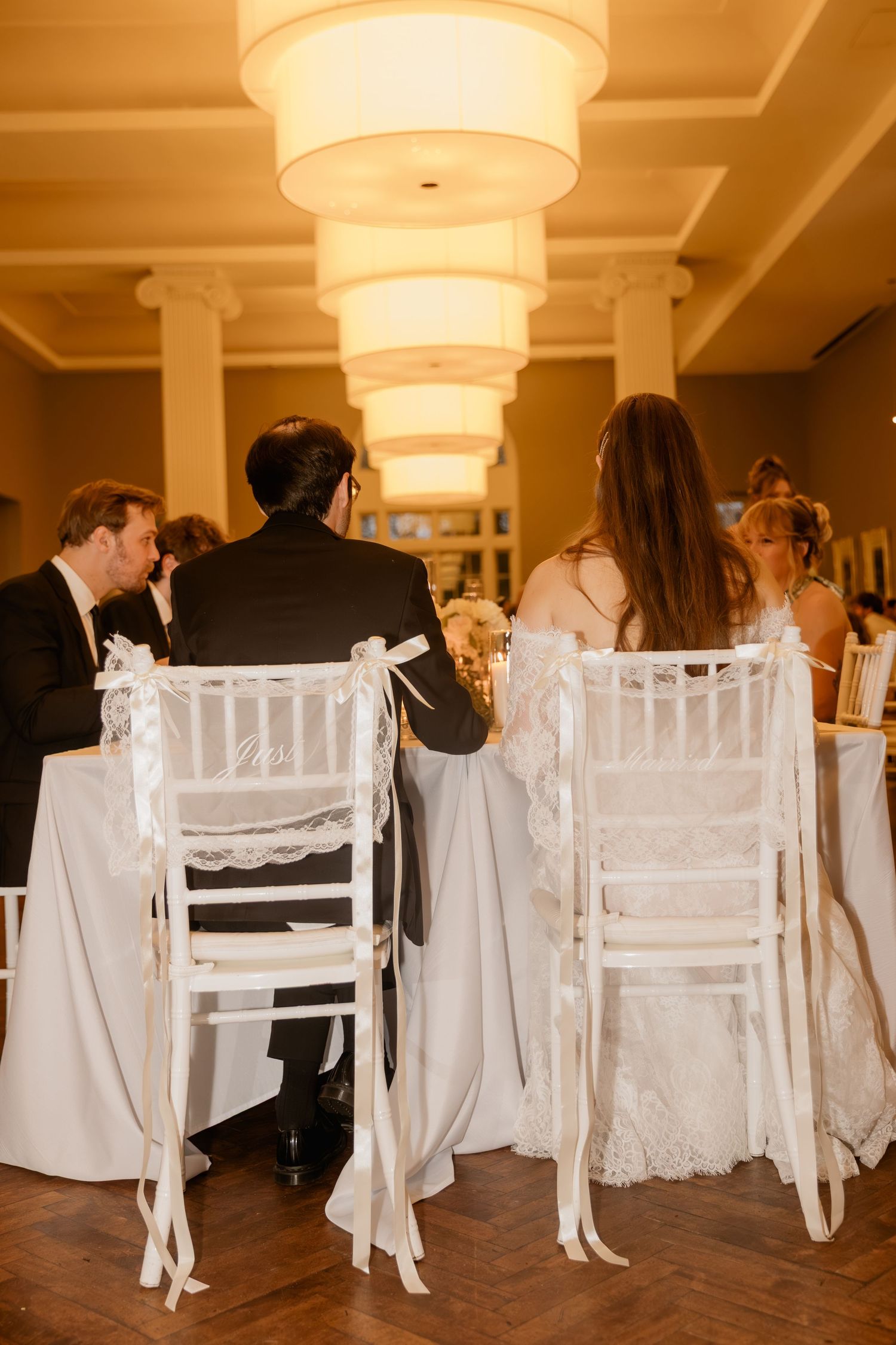 Bride and groom seated at wedding reception table with white chiavari chairs adorned with ribbons in elegant venue.