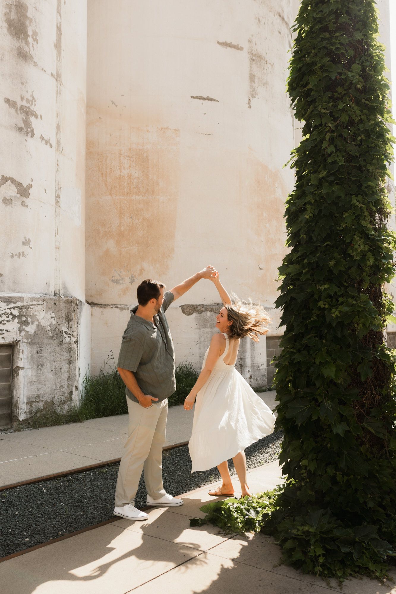 Couple dancing outdoors near ivy-covered column and rustic wall, woman in white dress twirling in sunlight.