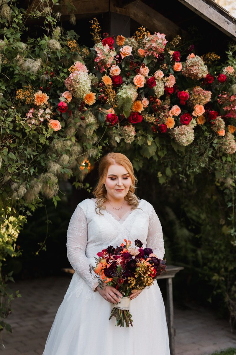 A white wedding gown adorned with floral arrangements featuring vibrant red and orange roses against a garden backdrop.
