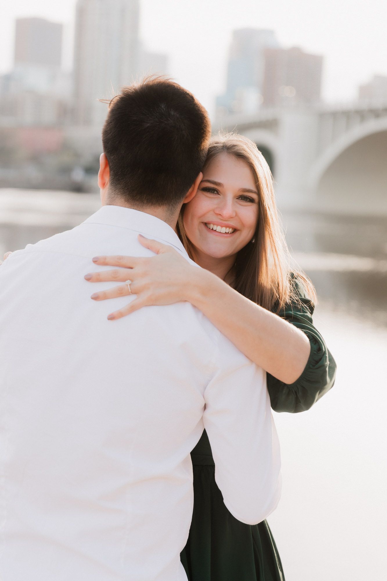 A smiling woman in a green dress hugs her partner near a river with a city bridge in the background.