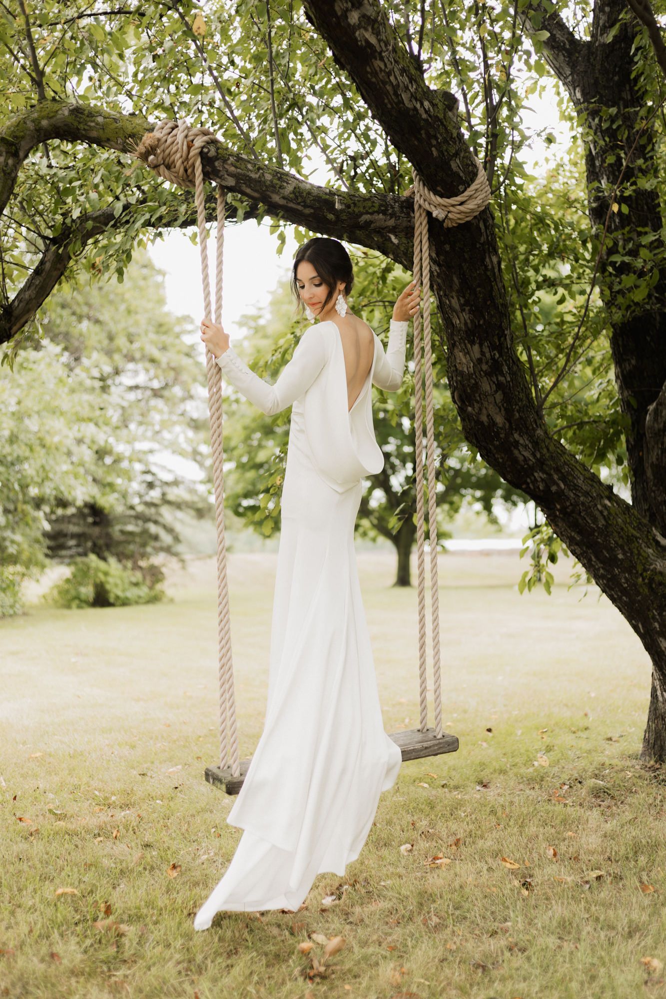 Bride in elegant white backless gown standing on rope swing under lush green tree in outdoor garden setting.
