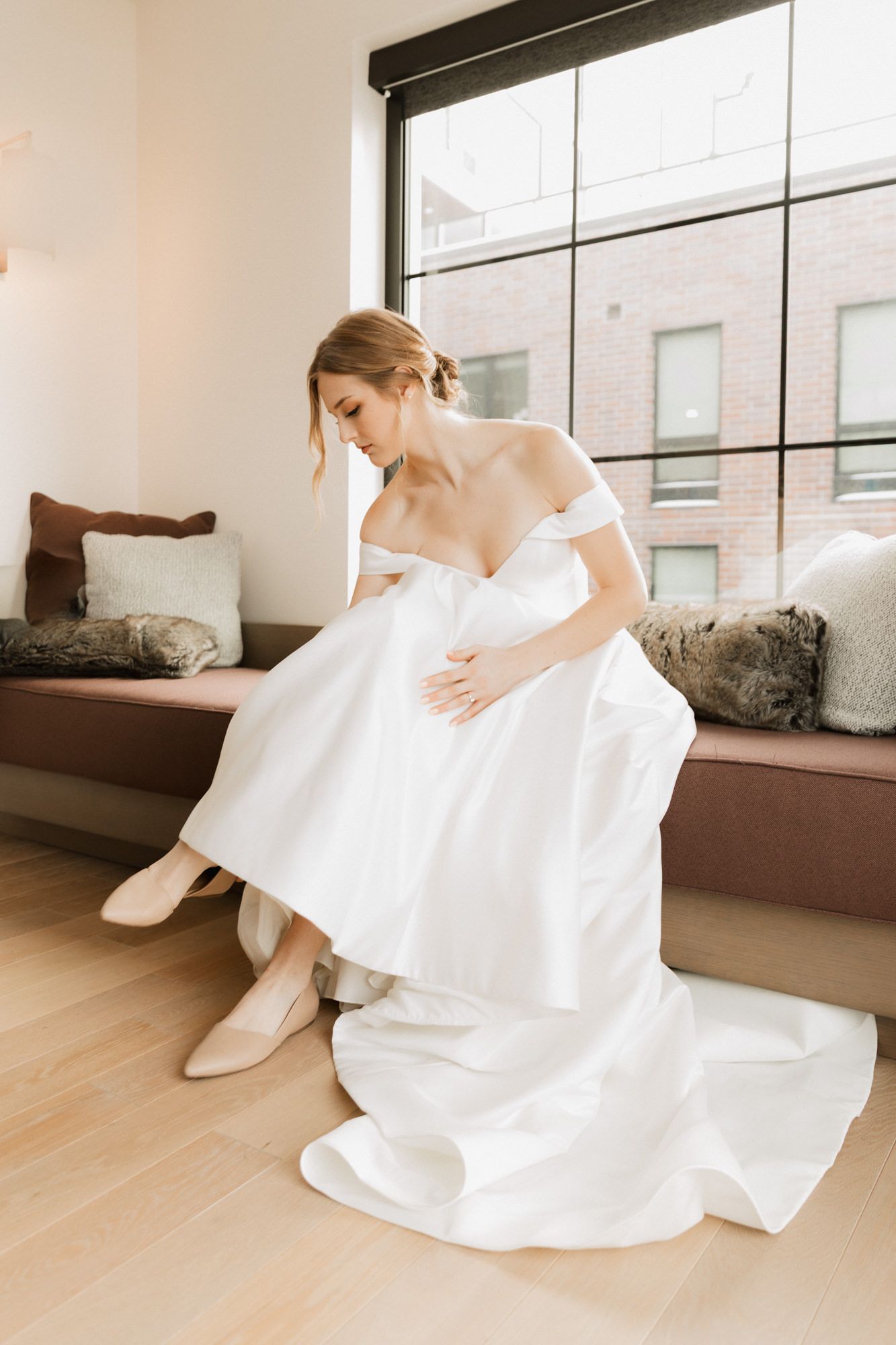Bride in off-shoulder white ball gown sitting by window, adjusting nude heels on wedding day.
