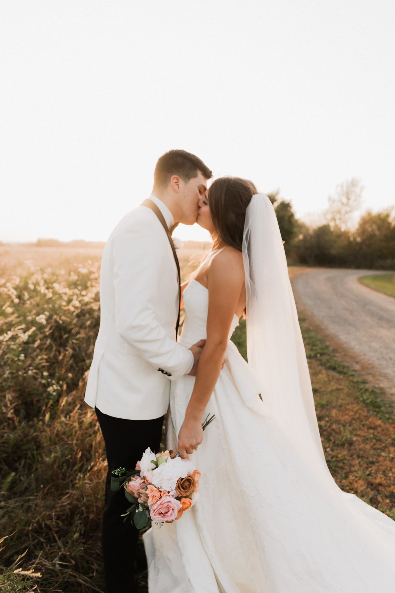 Bride and groom kissing at sunset in a field, bride holding a pink floral bouquet with veil flowing.