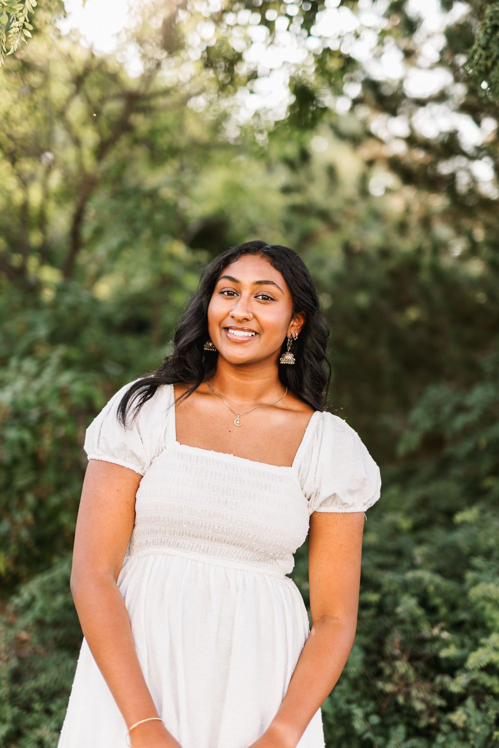 Outdoor portrait of person in white dress smiling against green foliage background.