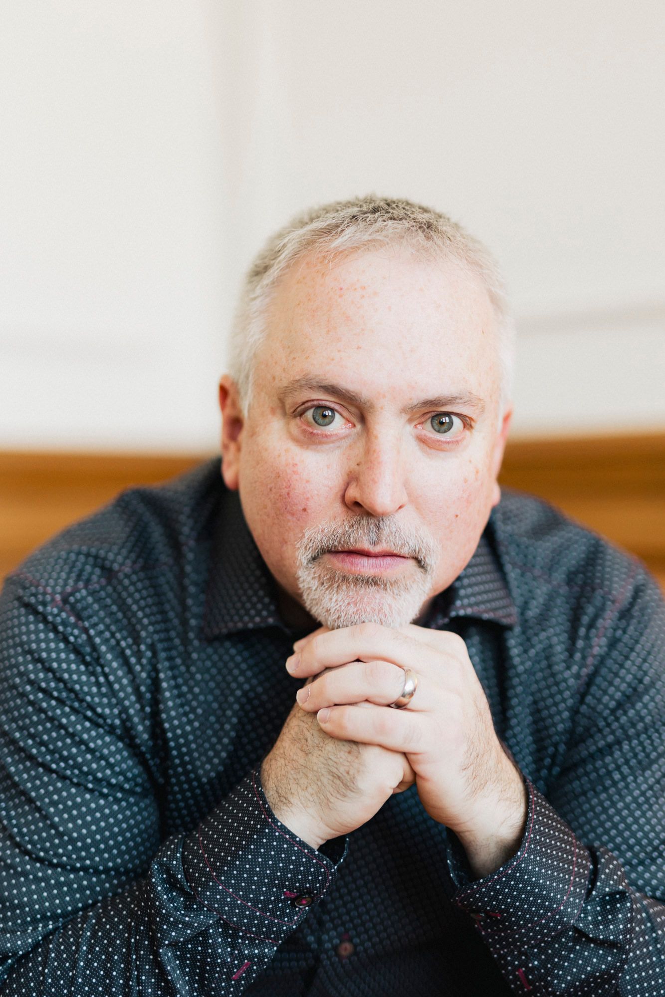 Professional headshot of person in patterned shirt with clasped hands in contemplative pose.